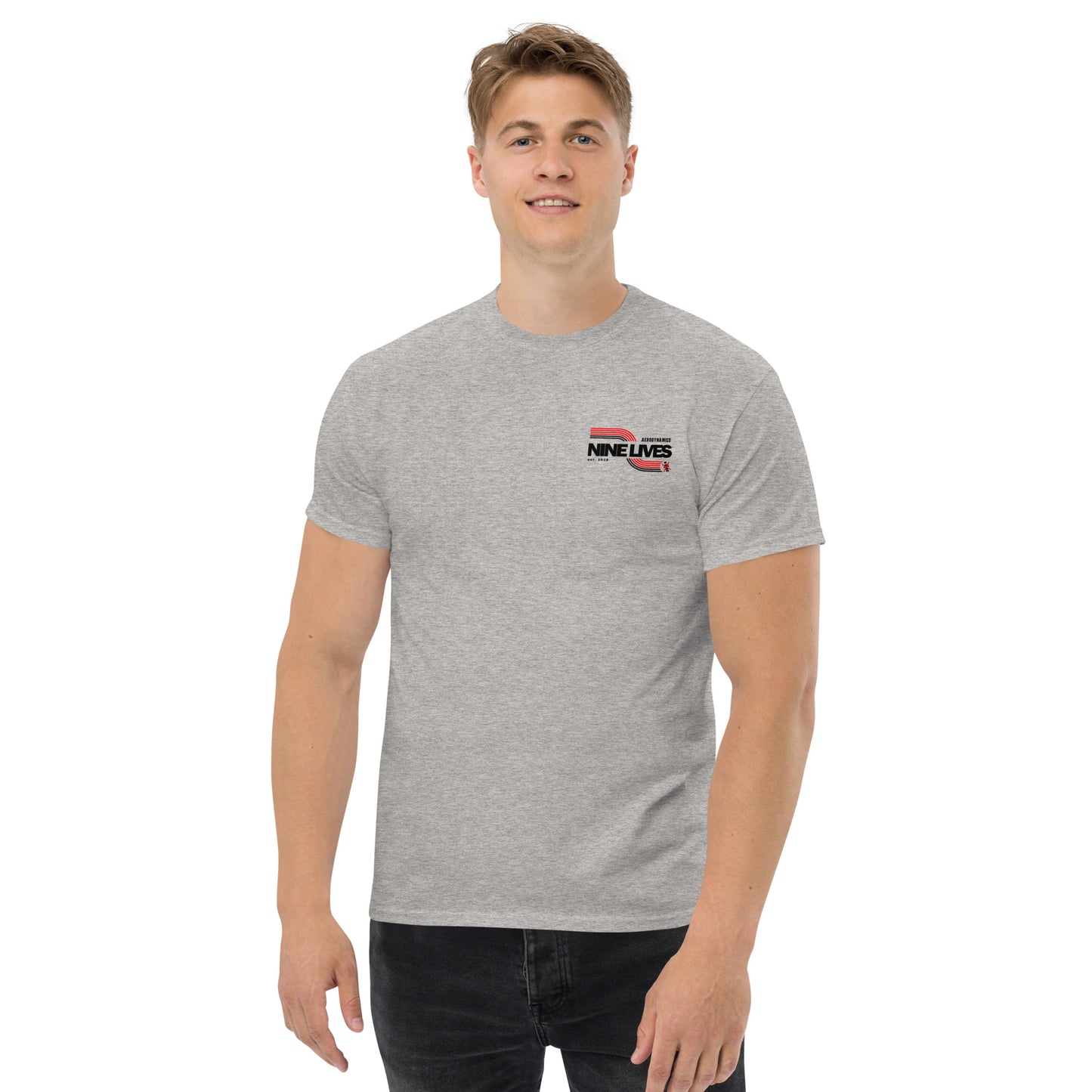 A smiling man with short, light brown hair is standing against a plain white background. He is wearing a grey Men's Soda Can Wang t-shirt from Nine Lives Racing made from 100% cotton featuring the words "Nine Lives" and a small graphic on the left side of his chest, paired with dark pants.