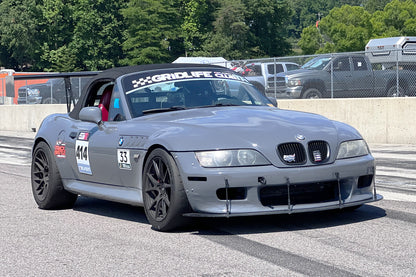 A gray BMW z3 roadster with the number 14 on the driver's door and 'Gridlife' branding on the windshield, sporting 9livesracing wings for improved lift-to-drag ratio and Big Wang GT3 performance modifications, speeds down a track with trees in the background and a barrier separating it from parked vehicles.