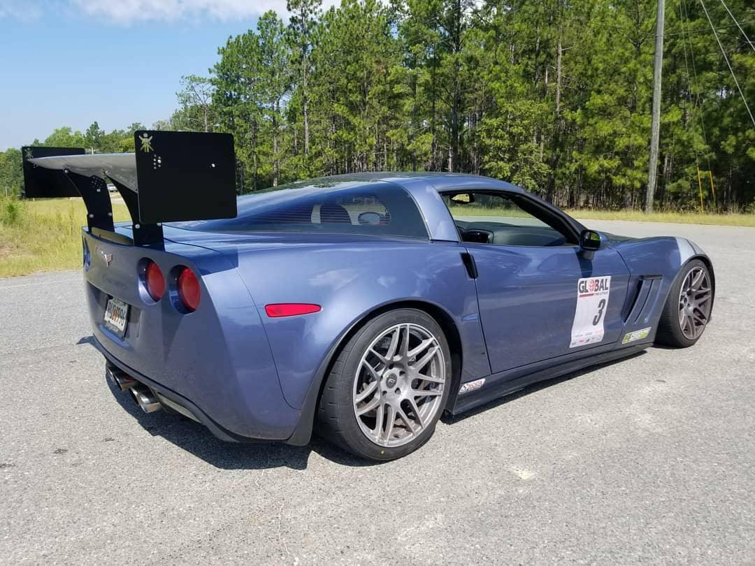 A blue sports car equipped with a 9livesracing Corvette Medium Downforce Kit ‘97-04 C5 is parked on an asphalt surface, with trees in the background. The car features sleek design lines, silver alloy wheels, and a racing sticker on the side displaying the number 3.