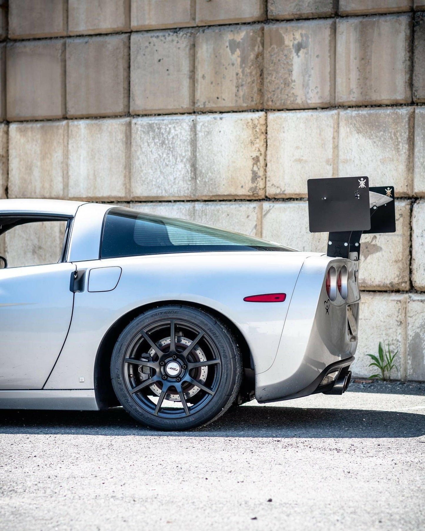 Side view of a silver C5 Corvette parked against an industrial concrete wall. The car features 9livesracing's Corvette Medium Downforce Kit ‘97-04 C5, which includes a large rear wing spoiler and dark alloy wheels designed to enhance downforce. The rear left corner and tail lights are visible, along with part of the driver's side window.