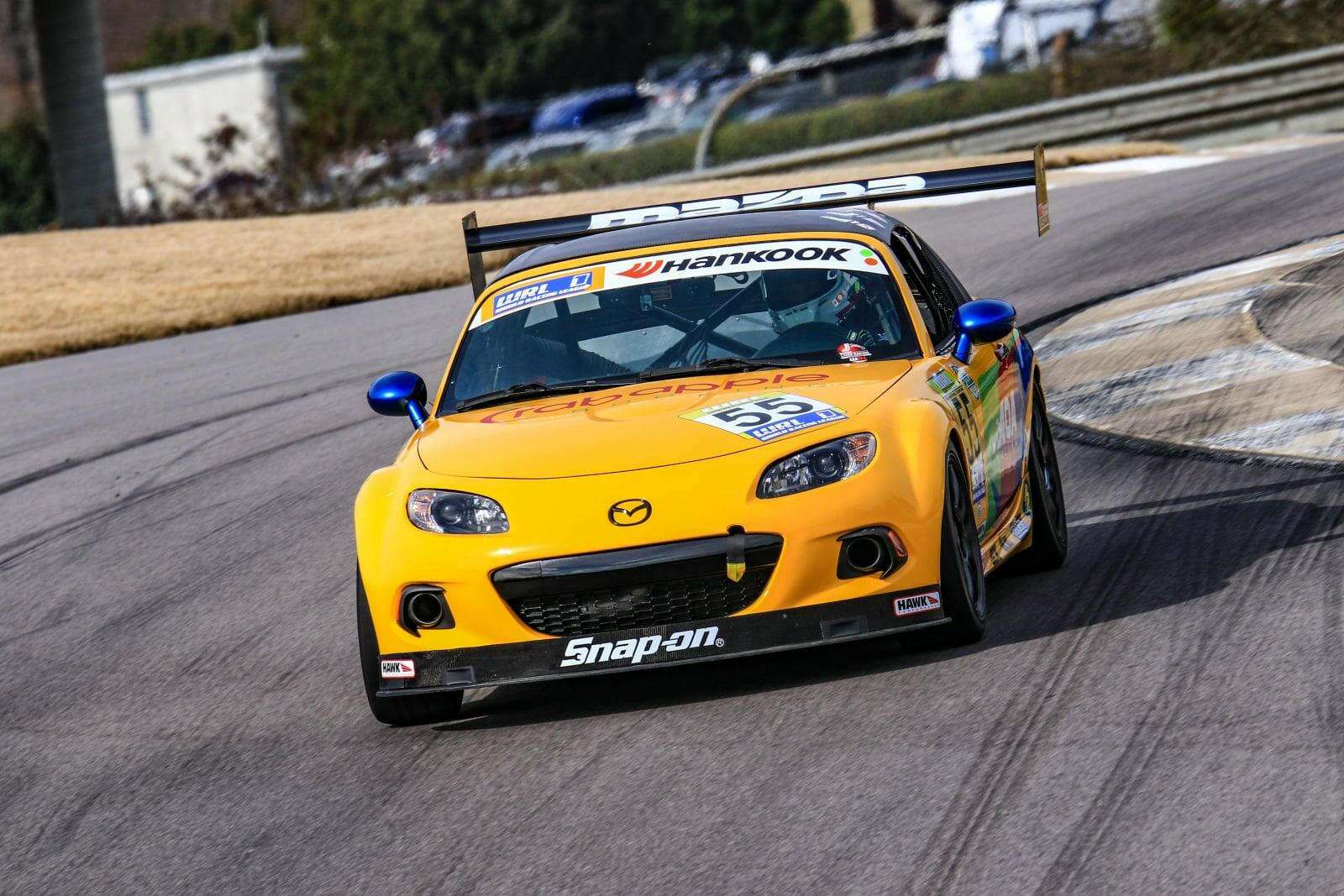 A yellow Mazda MX5 Splitters 06-15 NC, branded by 9livesracing and equipped with a Rocket Nose V2, speeds along a curved track. The car sports various sponsor decals, including Snap-on and Hankook. The driver is not visible in the image, and the background features a grassy area as well as several buildings.