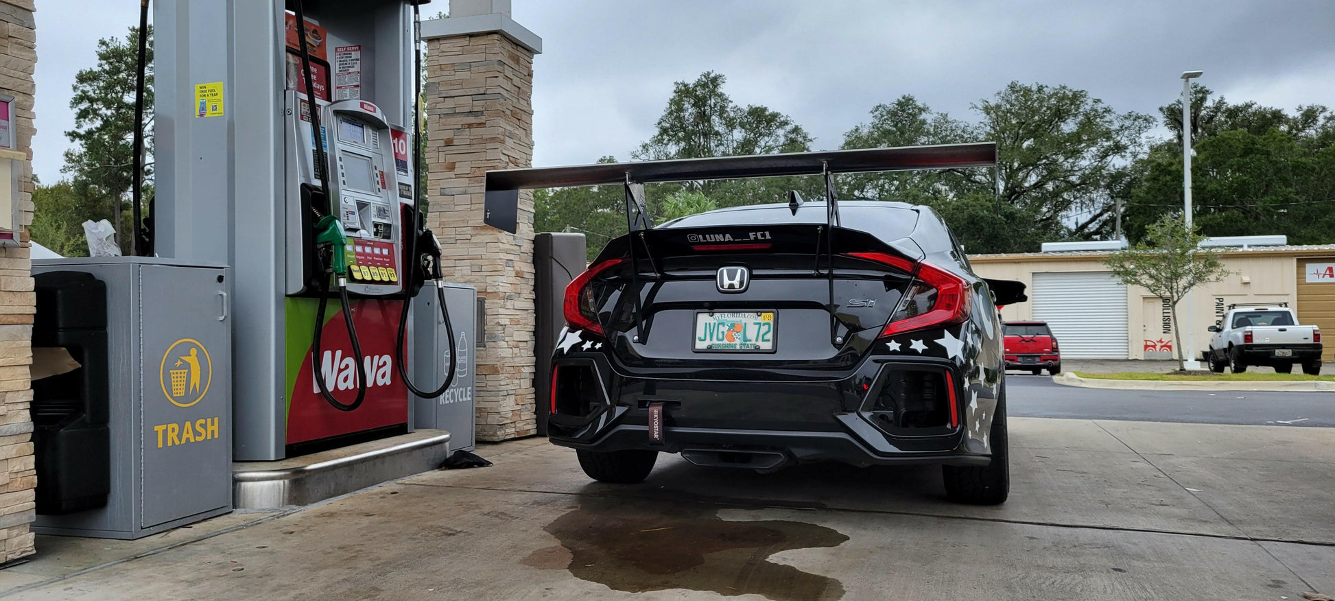 A black Honda Civic fitted with the 9livesracing Civic Wang kit 2017+ FC1 and sporting Florida license plates refuels at a Wawa gas station. The car's rear bumper is adorned with star decals, enhancing its already sporty appearance. Nearby, the gas pump island includes a trash bin, and in the background, there are trees, a 7-Eleven store, and a red vehicle.