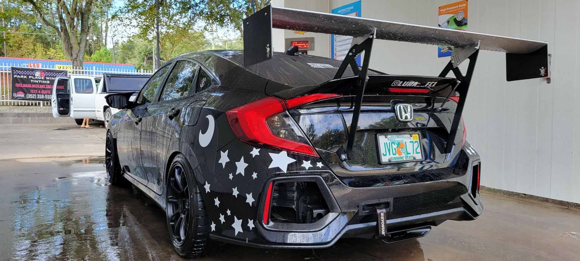 A black Honda adorned with star and moon decals on the rear and sides is parked inside a car wash. The vehicle boasts a Civic Wang kit 2017+ FC1 from 9livesracing, enhancing its downforce with a Big Wang GT3 spoiler, and also features custom license plates. The ground in the car wash area is wet, while trees can be seen in the background outside the wash bay.