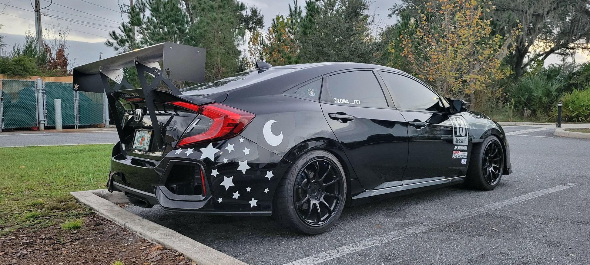 A black car with black rims is parked at an angle in a parking lot. The car features white star and moon decals on the sides and rear, a Civic Wang kit 2017+ FC1 by 9livesracing for optimal downforce, and various performance stickers. Trees and a fence are visible in the background.