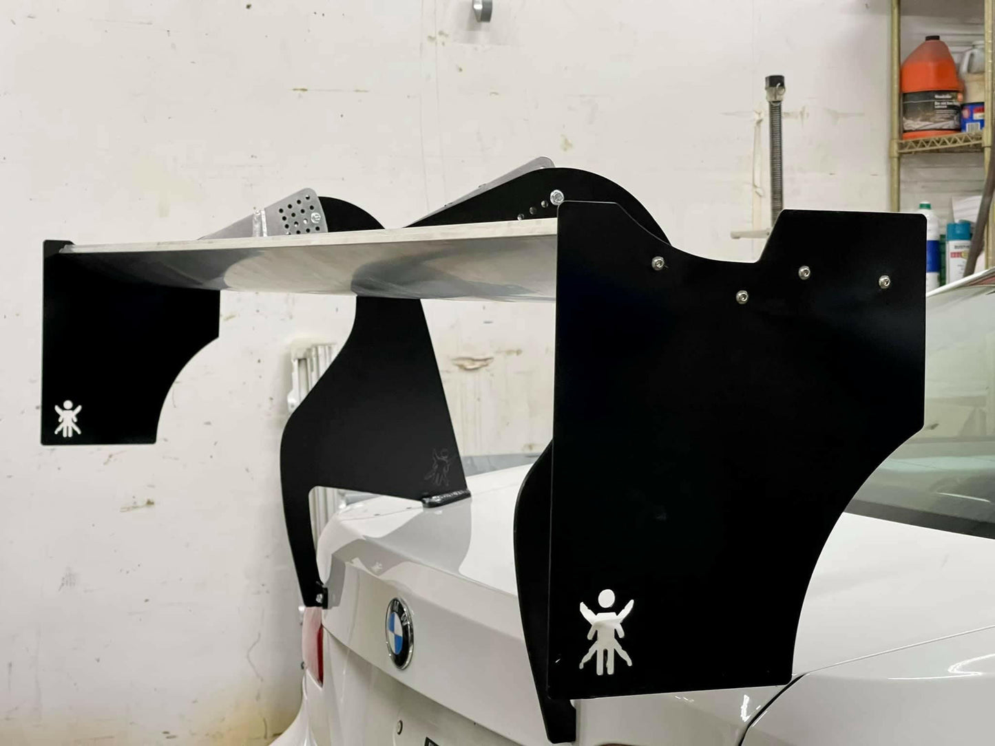 A large spoiler is mounted on the rear of a white BMW car. The spoiler features black End Plates for The Big Wáng! by 9livesracing, with a logo of a stick figure holding up its arms. Engineered for optimal downforce, the aluminum setup complements the garage backdrop filled with tools and equipment.