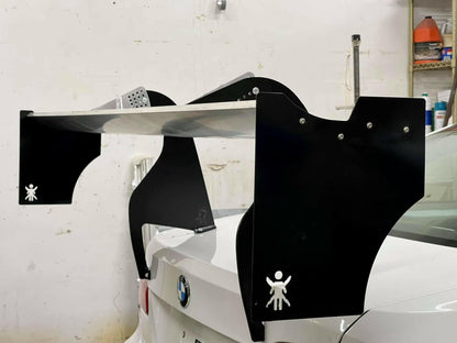 A large spoiler is mounted on the rear of a white BMW car. The spoiler features black End Plates for The Big Wáng! by 9livesracing, with a logo of a stick figure holding up its arms. Engineered for optimal downforce, the aluminum setup complements the garage backdrop filled with tools and equipment.