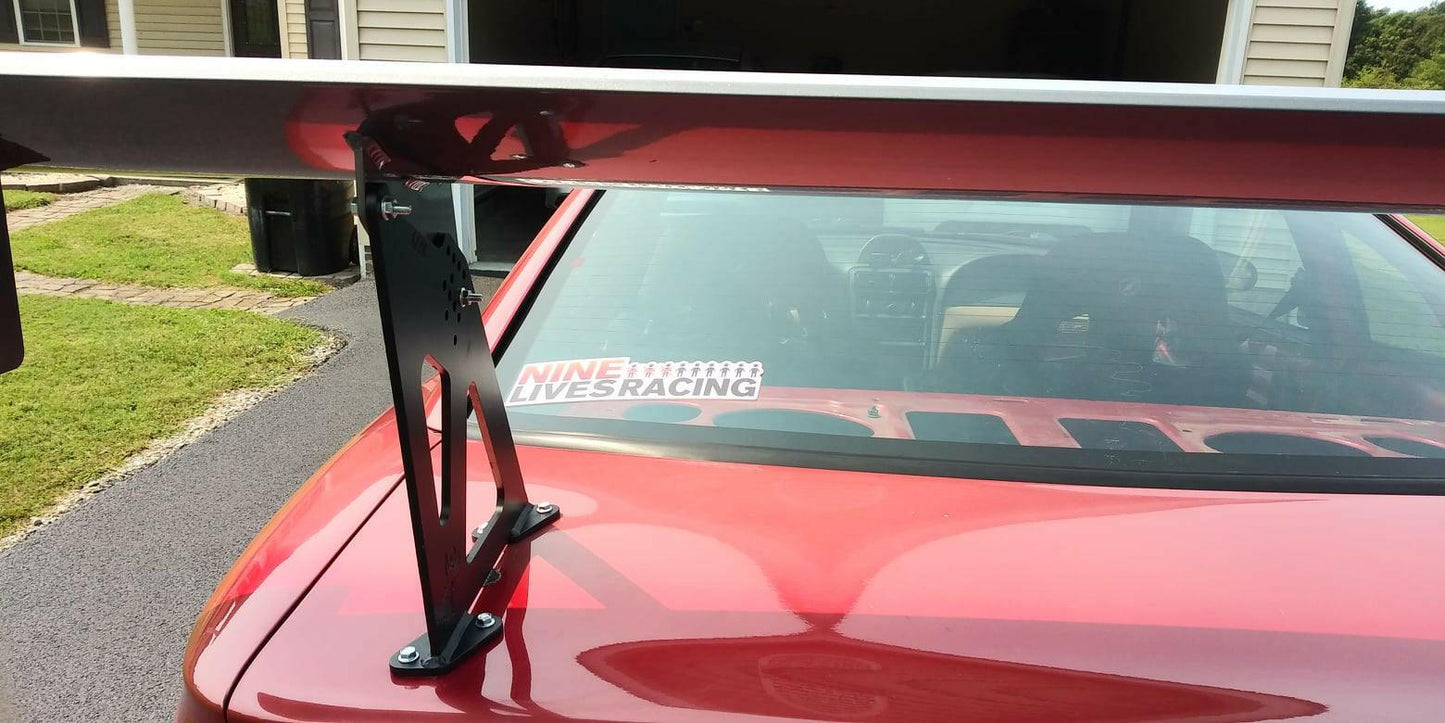 A close-up of the rear section of a red car, showcasing a Mustang Big Wang Kit '94-‘98 SN95 rear wing spoiler mounted on the trunk. The spoiler features extruded aluminum wings and is supported by black brackets, enhancing its lift-to-drag ratio. A "9livesracing" decal is visible on the back window. In the background, part of a garage and yard can be seen.
