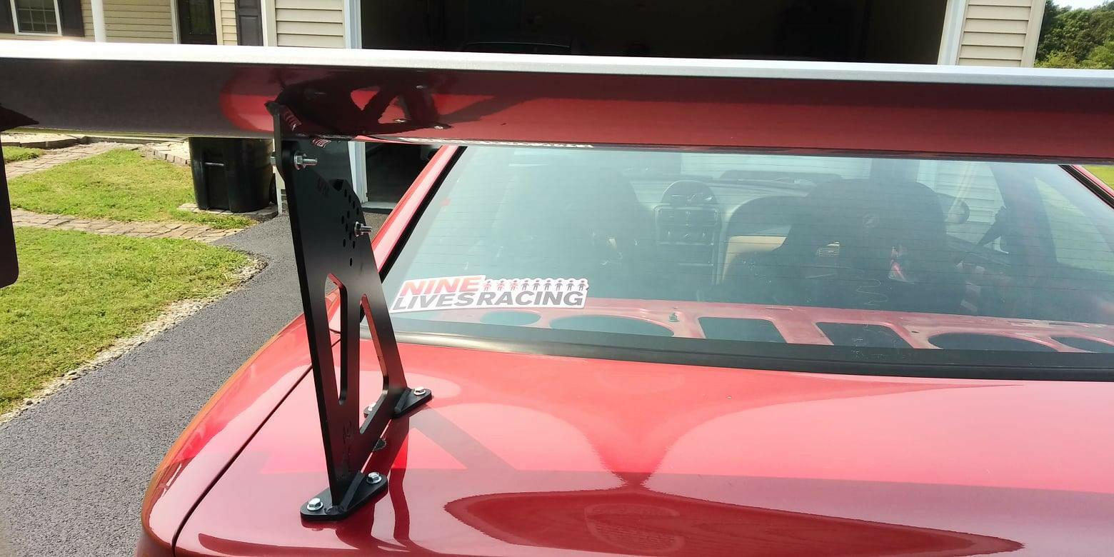 A close-up of the rear section of a red car, showcasing a Mustang Big Wang Kit '94-‘98 SN95 rear wing spoiler mounted on the trunk. The spoiler features extruded aluminum wings and is supported by black brackets, enhancing its lift-to-drag ratio. A "9livesracing" decal is visible on the back window. In the background, part of a garage and yard can be seen.