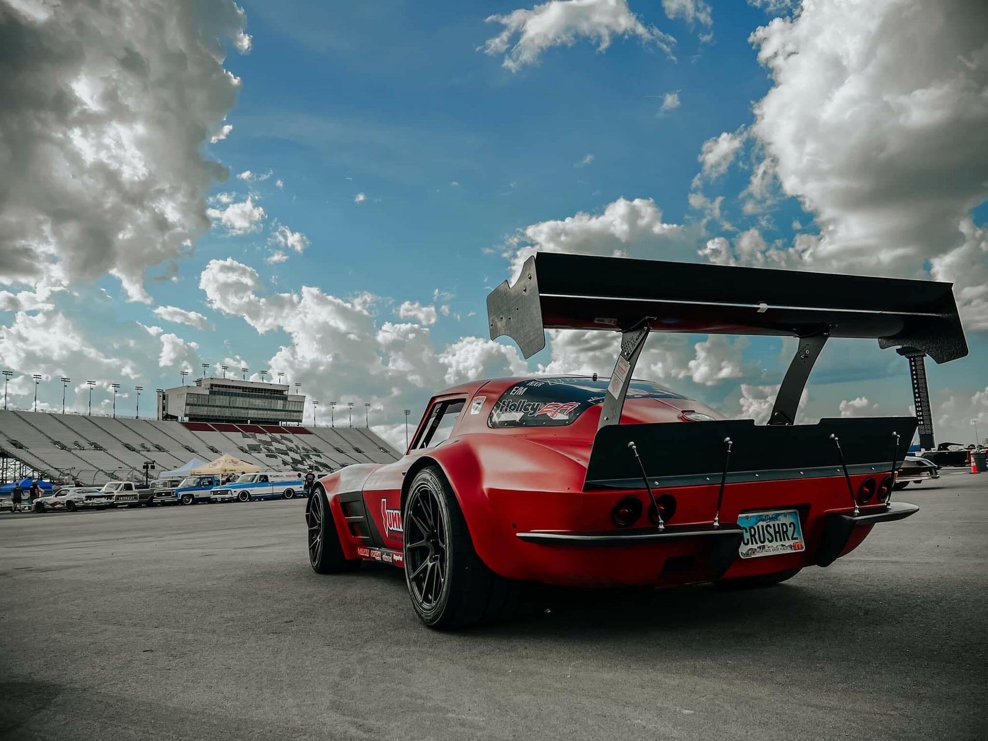 A low-angle shot of a red race car featuring "The Deuce! Carbon Double Element Kit" by 9livesracing, parked on a track. The car has a "RUSH2R" license plate. The sky above is partly cloudy, with bleachers and other vehicles visible in the background.