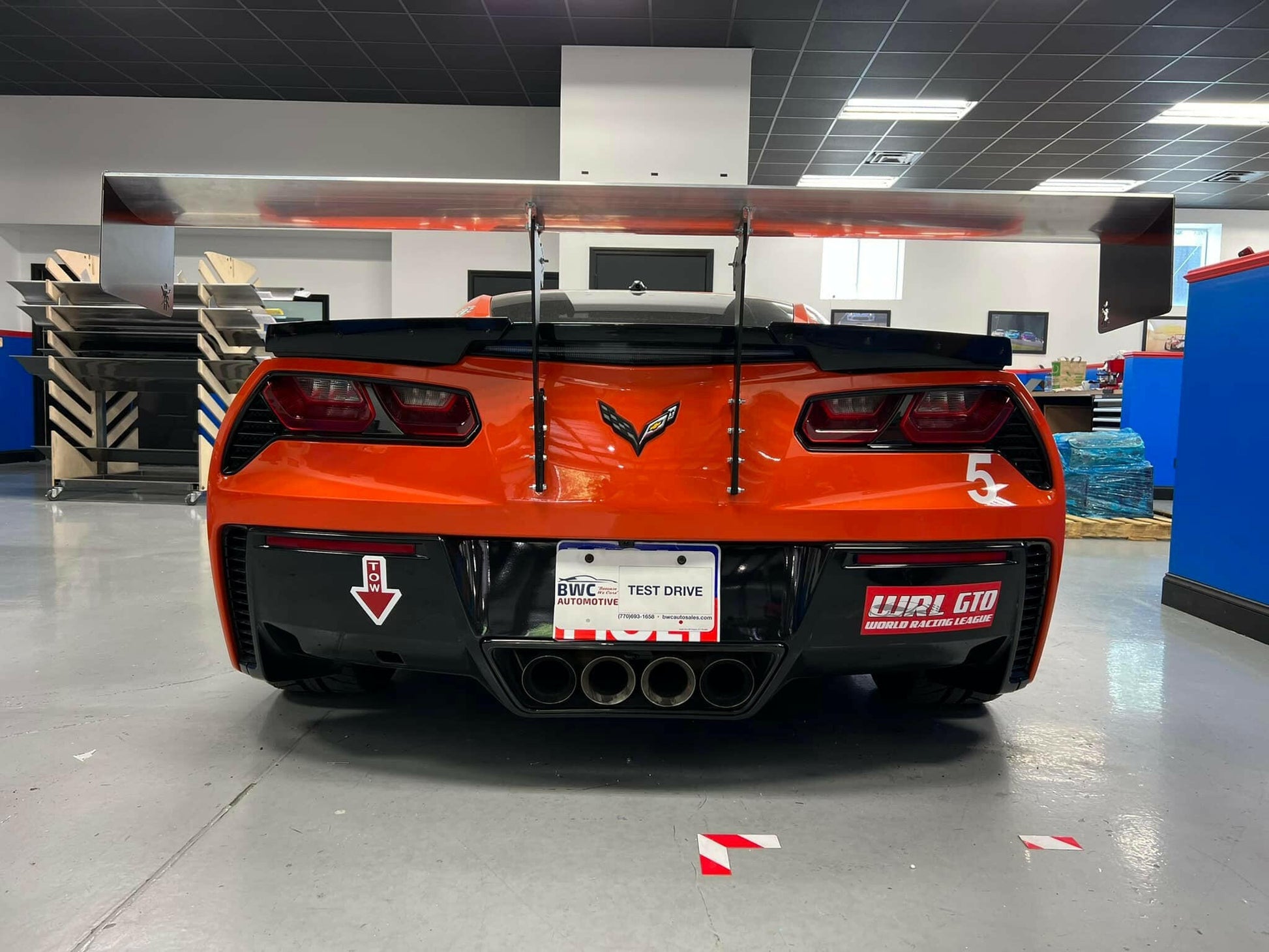 A red Corvette Big Wang Chassis Mount (2014-2019 C7) by 9livesracing, featuring a large rear spoiler with down-force adjustments, is shown from the back, parked indoors. The car boasts distinctive tail lights, four exhaust pipes, and decals including a white number 53 and a "Test Drive" license plate. The surrounding area appears to be a garage or workshop.