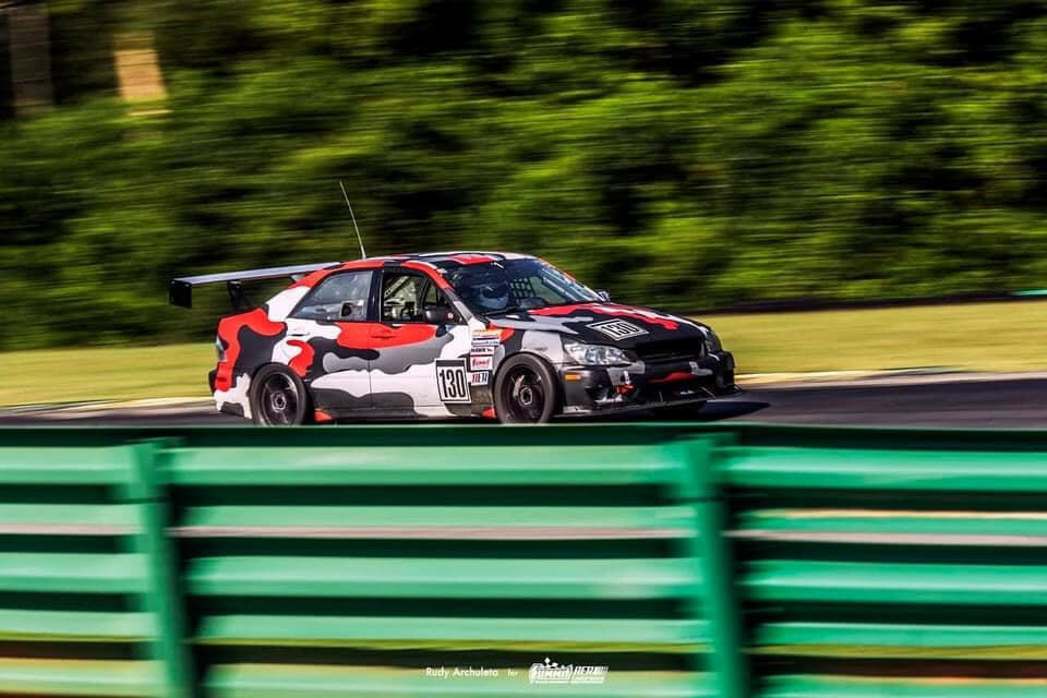 A car sporting the Is300 Big Wang Kit 98-04 xe10 from 9livesracing, adorned in a red, black, and white camouflage paint job and numbered 130, races on the track. It showcases a large rear wing engineered for optimal down-force adjustments and is covered in multiple sponsor decals. Green barriers line the track while the blurred background indicates high speed.
