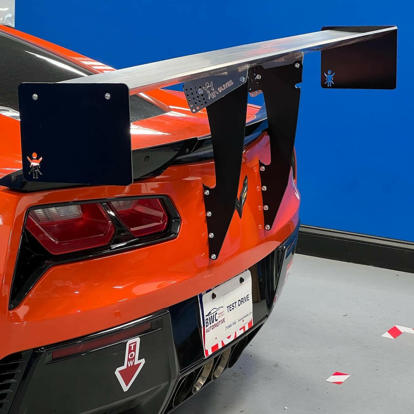 A bright orange sports car with a large custom rear wing, specifically the Corvette Big Wang Chassis Mount ’2014 - 2019 C7 by 9livesracing, attached to the back. The wing has black end plates adorned with small stickers depicting a waving stick figure. The car sports a "TEST DRIVE" license plate and a tow hook indicator sticker on the lower rear bumper.