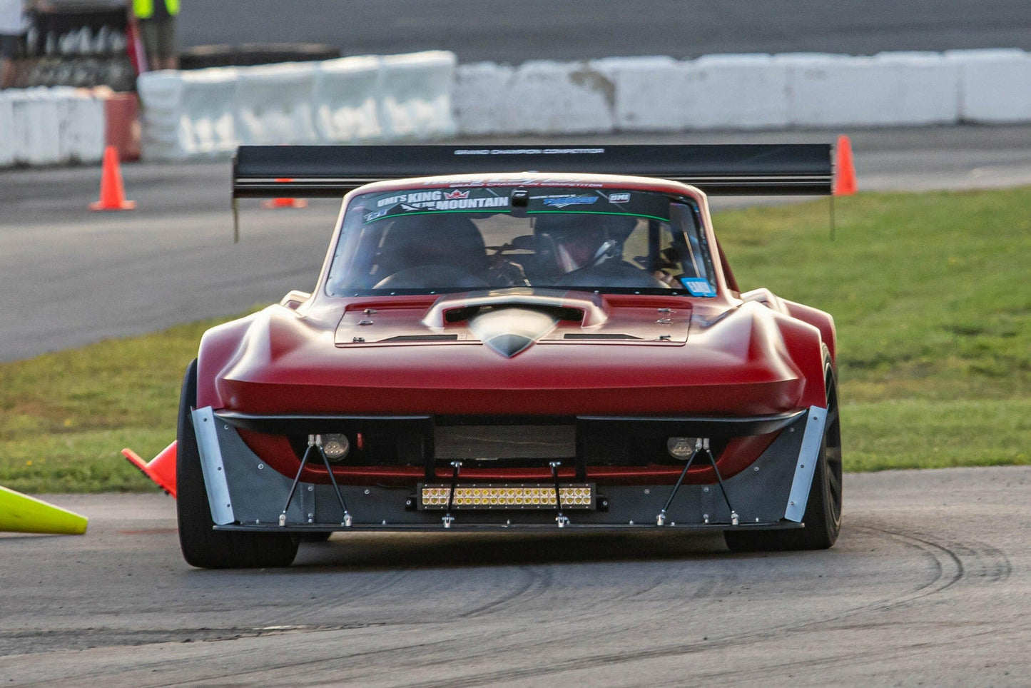 A maroon race car equipped with the wide body kit and a sizable carbon wing from 9livesracing's "The Deuce! Carbon Double Element Kit" is captured in motion on a race track, skillfully navigating around orange traffic cones. The car boasts aggressive aerodynamic features and meticulous modifications, showcasing its design for high-performance racing.