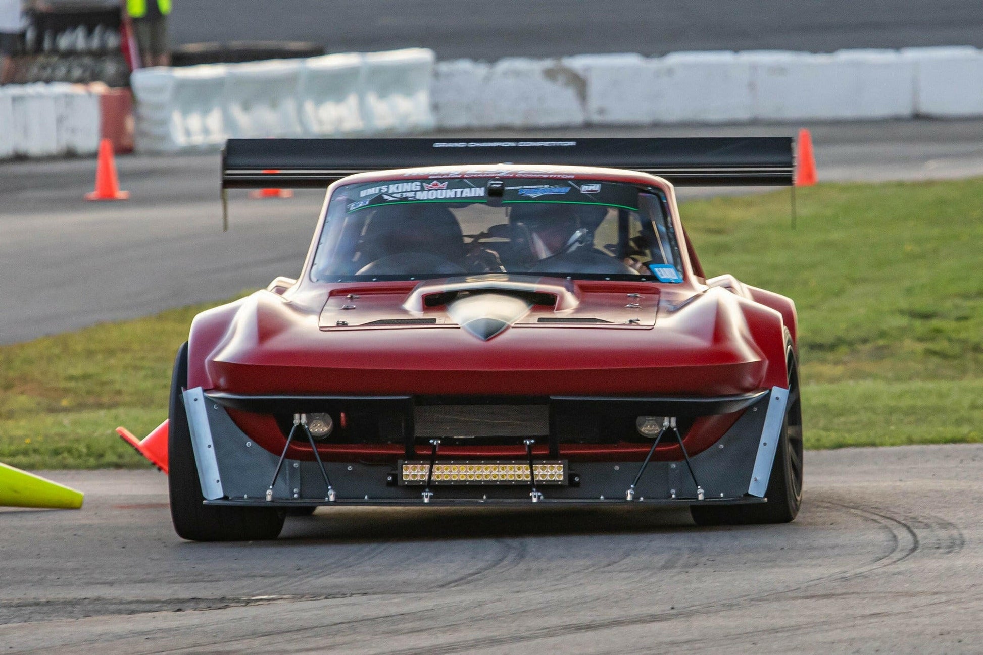 A maroon race car equipped with the wide body kit and a sizable carbon wing from 9livesracing's "The Deuce! Carbon Double Element Kit" is captured in motion on a race track, skillfully navigating around orange traffic cones. The car boasts aggressive aerodynamic features and meticulous modifications, showcasing its design for high-performance racing.