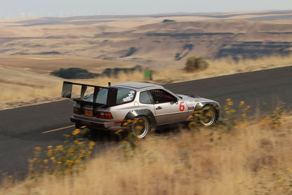 A silver Porsche 944 on a canyon road featuring a 9 lives racing big wing.