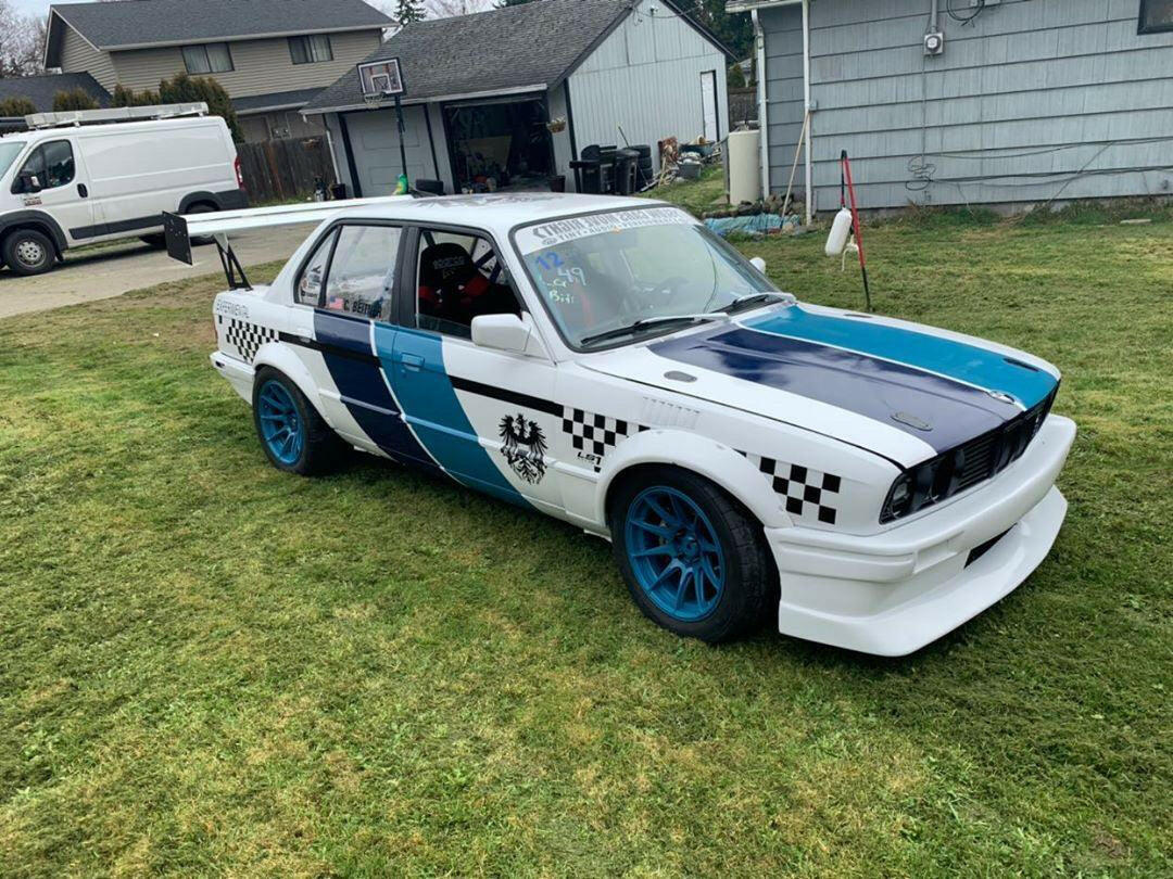 A white E30 BMW sedan, equipped with the 325 Big Wang kit ’82-94 E30 from 9livesracing, is parked on grass. The car features a blue racing stripe, blue and black checkered pattern, and blue wheels. It has various decals, a rear spoiler for added downforce, and a front splitter. In the background, there is a white van and residential houses.