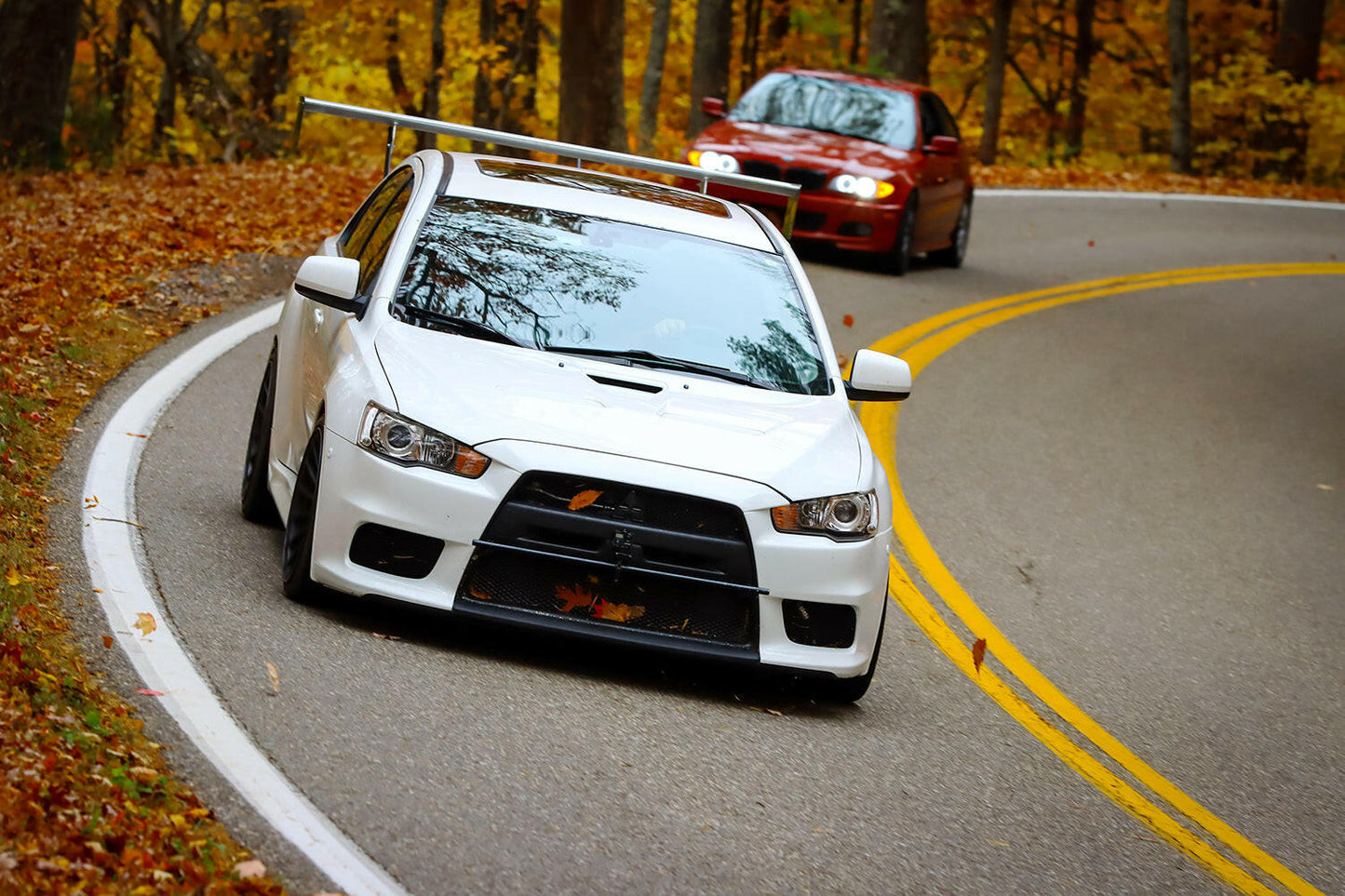 A white sports car equipped with the 9livesracing Evolution 10 Big Wang kit, complete with aluminum wings and a large rear spoiler, and adorned with an orange leaf on its front grill, drives around a winding road lined with autumn leaves. Closely behind it follows a red car. The trees on either side showcase fall foliage in shades of yellow, orange, and brown.