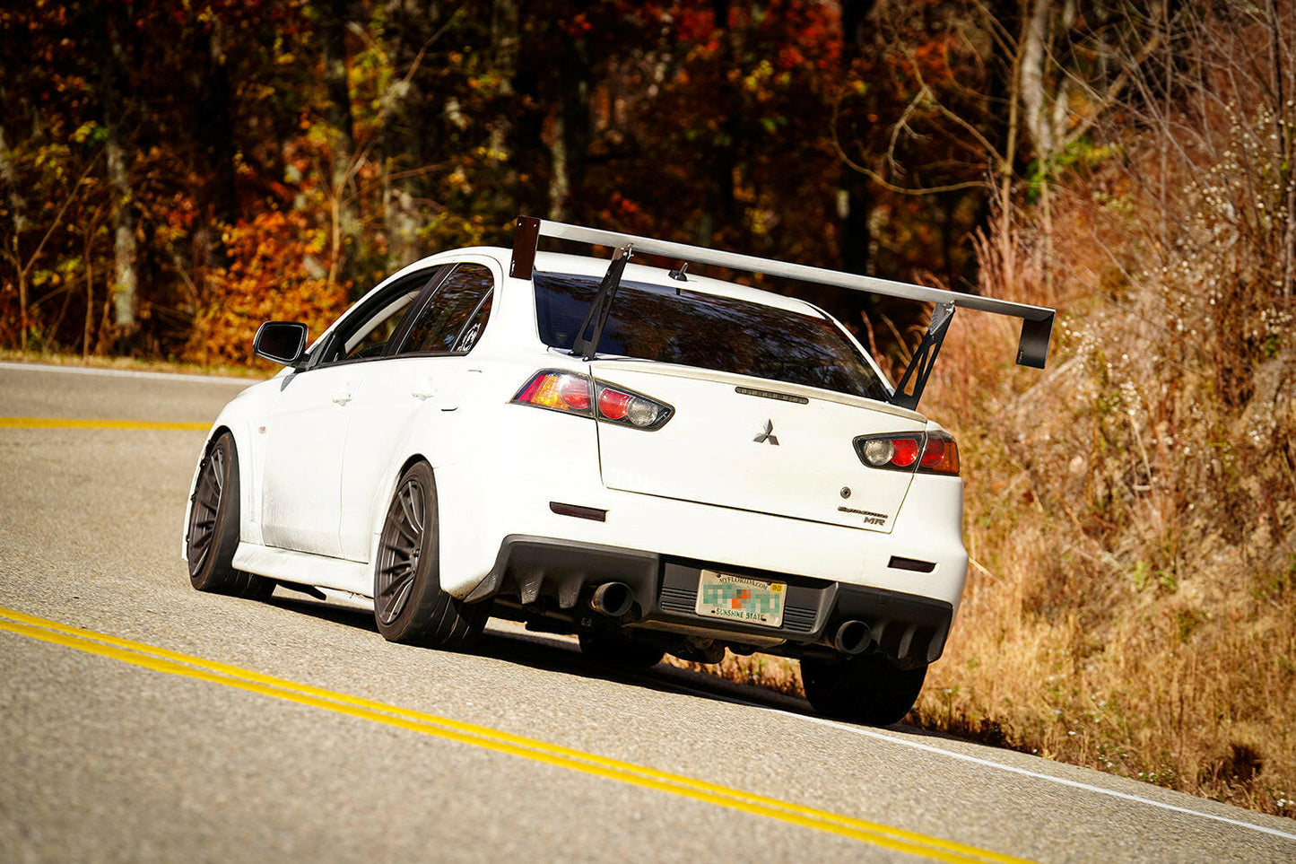 A white Mitsubishi Lancer Evolution, equipped with the Evolution 10 Big Wang kit from 9livesracing, gracefully navigates a winding road flanked by autumn-colored trees. The car boasts a prominent rear spoiler and aluminum wings, along with black rims and tinted windows, capturing the motion and vibrant fall foliage beautifully.