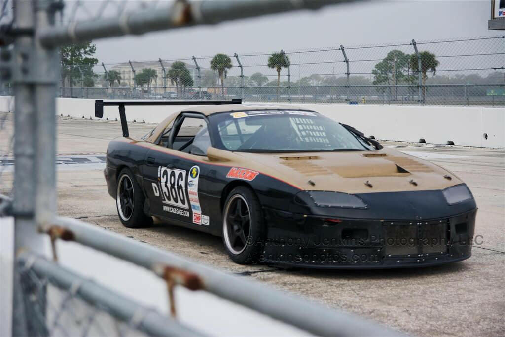 A black and bronze race car with the number 386 on the side is parked on a race track. Adorned with sponsor decals and a large rear spoiler mounted using the Universal DIY Mounts - Pylons Only by 9livesracing, it cuts an impressive figure against the chain-link fence and palm trees. The ambiance is overcast with a cloudy sky.