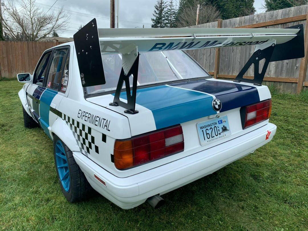 Rear view of an E30 BMW race car with blue accents, equipped with the 325 Big Wang kit ’82-94 E30 from 9livesracing for optimal downforce. The car showcases a checkered flag pattern and "EXPERIMENTAL" on the side. The license plate reads "1620X," and the vehicle is parked on grass near a wooden fence.