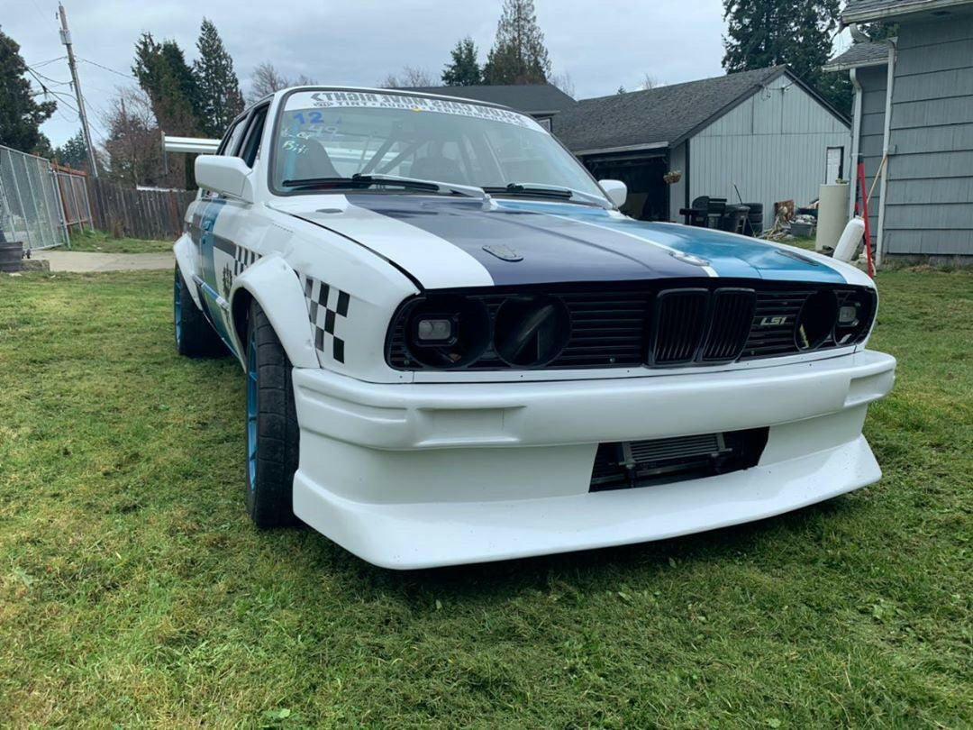 A white and blue E30 BMW, equipped with a wide body kit and checkered pattern details, is parked on a grassy lawn. The car features blacked-out headlights, a front bumper with a prominent air intake, and the 325 Big Wang kit ’82-94 E30 from 9livesracing for added downforce. Buildings and trees can be seen in the background.