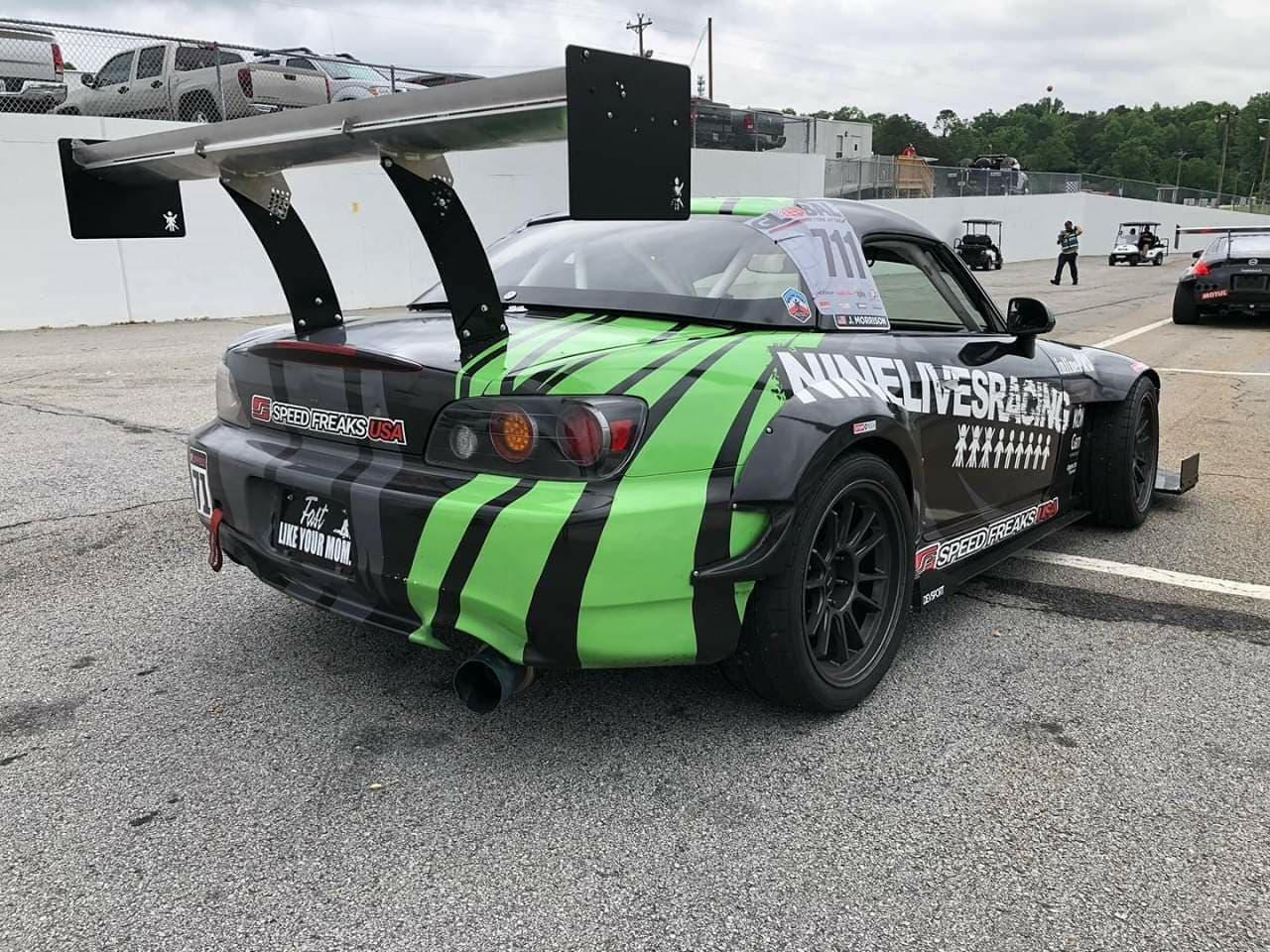 A race car showcasing a black and green striped design with branding for 9livesracing and Speed Freaks USA is parked in a lot. Equipped with The Deuce! Double Element Kit, the American-made car proudly displays the number 771 on its side window. Other vehicles and a white wall can be seen in the background.