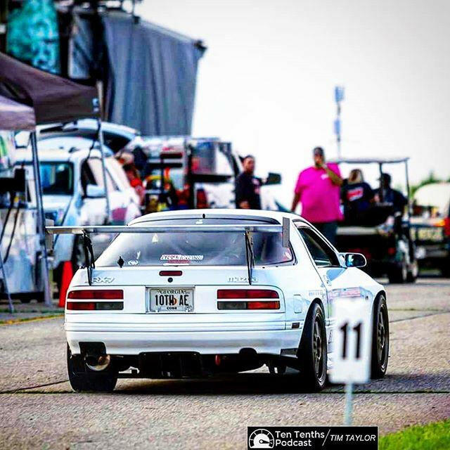 At an outdoor event, a white sports car featuring rear spoiler pylons from 9livesracing, aluminum mounts, and racing decals is parked. Resembling an FC Rx7, the car's license plate reads "10TH AE." The bustling background shows several people and cars. Photo credit: Ten Tenths / Tim Taylor Podcast.