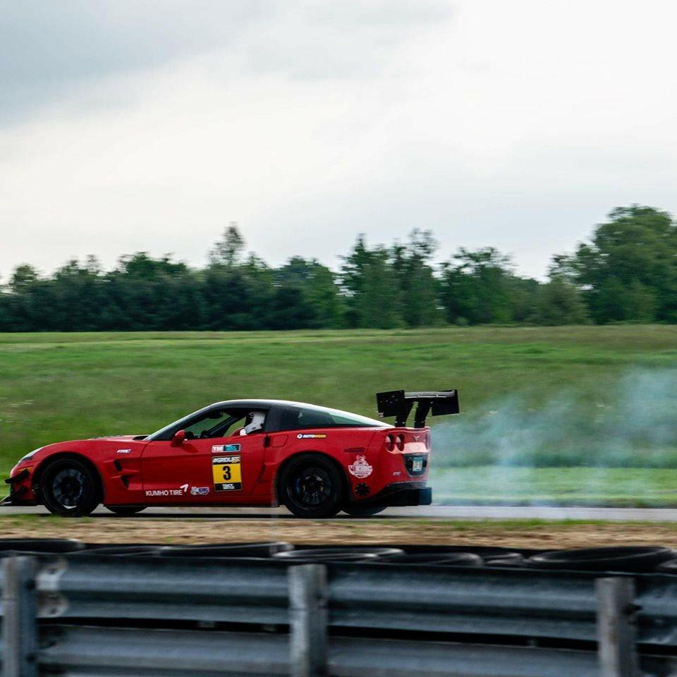 A red Corvette race car with the number 3 on its door drifts on a racetrack, producing smoke from its rear tires. Featuring 9livesracing’s Corvette Big Wang Chassis Mount ’97-13 C5/6 extruded aluminum wings, including a Big Wang GT3 rear wing, it boasts an optimized lift-to-drag ratio. The track is surrounded by green grass and trees beneath an overcast sky.