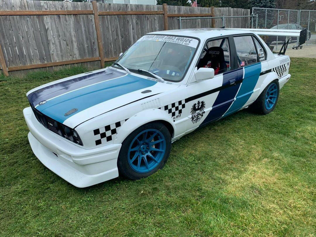 A white classic E30 BMW with blue accents and a racing number on the door, featuring a distinctive checkerboard pattern on the sides, is parked on a grassy area near a wooden fence. The car boasts blue rims and sporty modifications like a body kit, rear spoiler, and the 325 Big Wang kit from 9livesracing for added downforce.