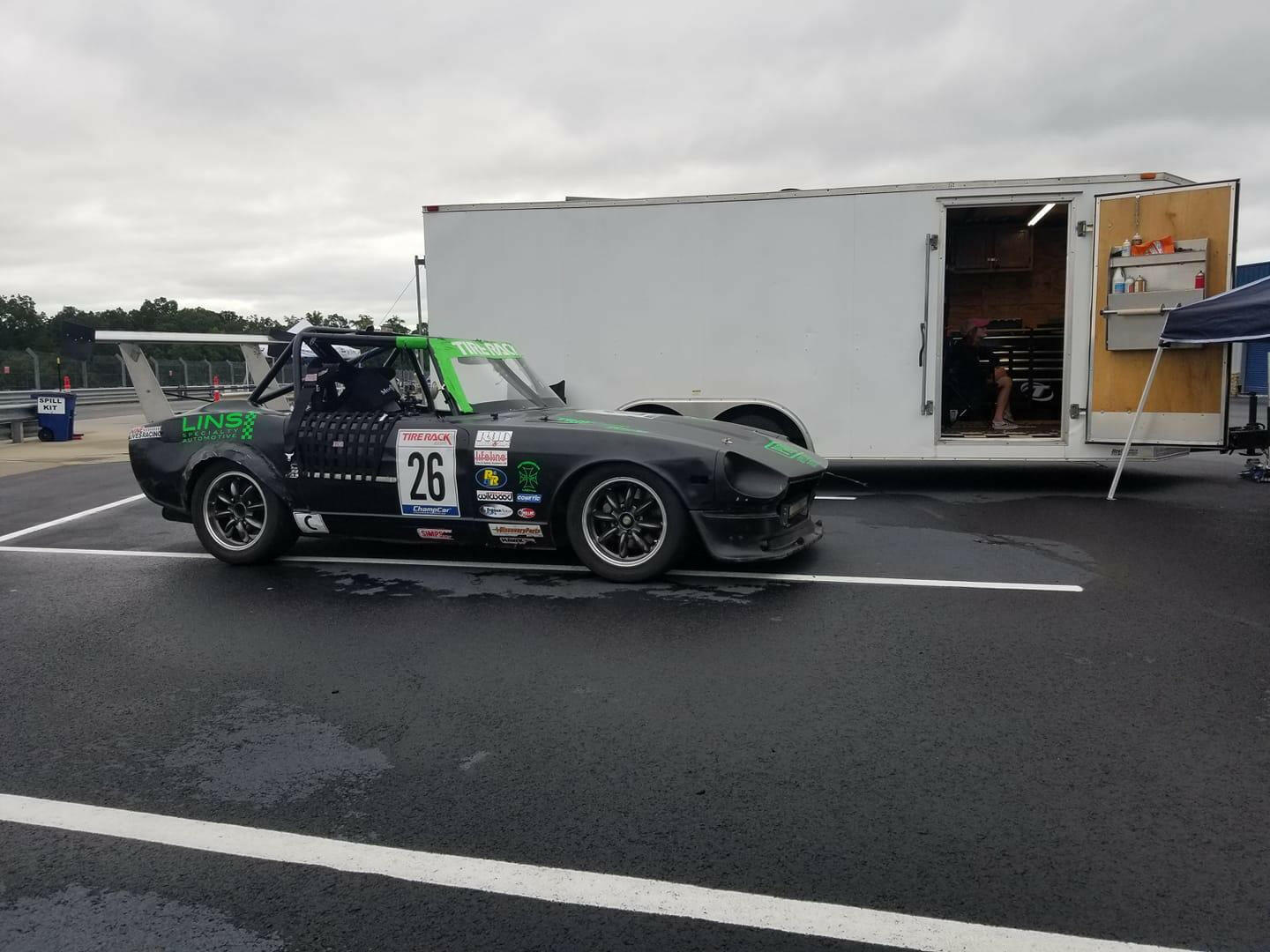 A sleek black race car with green accents, labeled number 26, is parked on an asphalt track next to a large white trailer. The cloudy sky and trees are visible in the background. Sporting 9livesracing Universal DIY Mounts - Pylons Only and various sponsorship logos, this machine is built for speed.