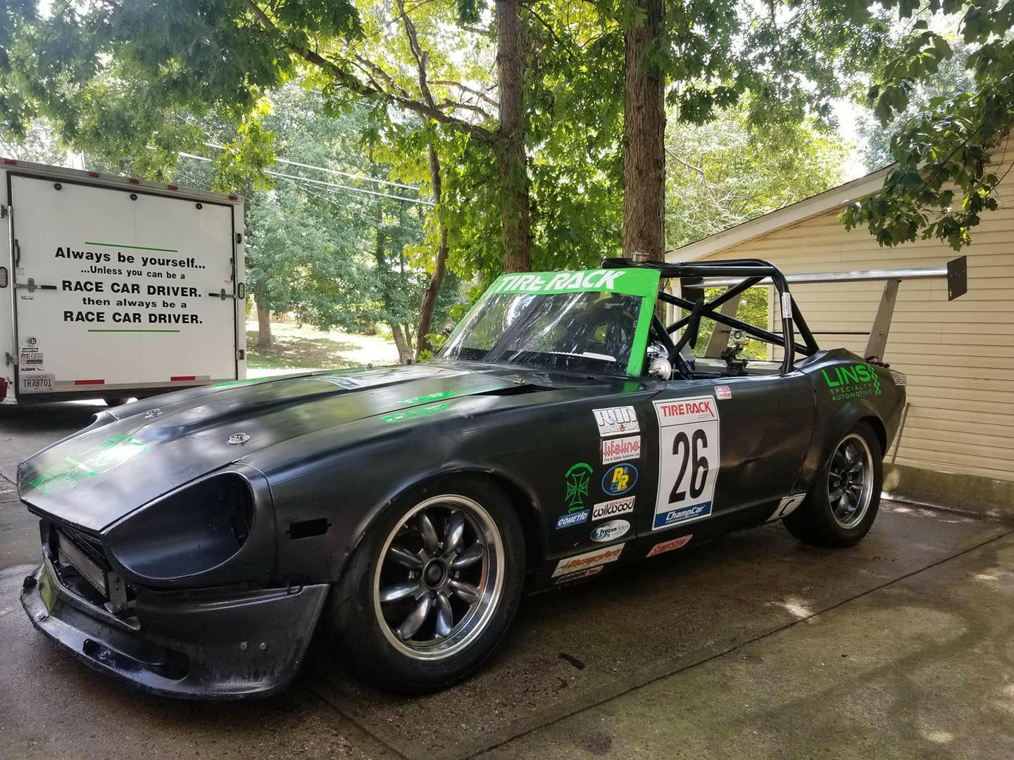 A black race car displaying the number 26 on the door sits in a driveway. The vehicle, adorned with sponsor decals such as "Tire Rack" and "Lins," is equipped with custom aluminum mounts from the Universal DIY Mounts - Pylons Only by 9livesracing. In the background, a sign reads, "Always be yourself... unless you can be a RACE CAR DRIVER." Trees and a house are visible nearby.