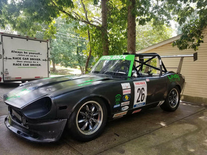 A black race car displaying the number 26 on the door sits in a driveway. The vehicle, adorned with sponsor decals such as "Tire Rack" and "Lins," is equipped with custom aluminum mounts from the Universal DIY Mounts - Pylons Only by 9livesracing. In the background, a sign reads, "Always be yourself... unless you can be a RACE CAR DRIVER." Trees and a house are visible nearby.