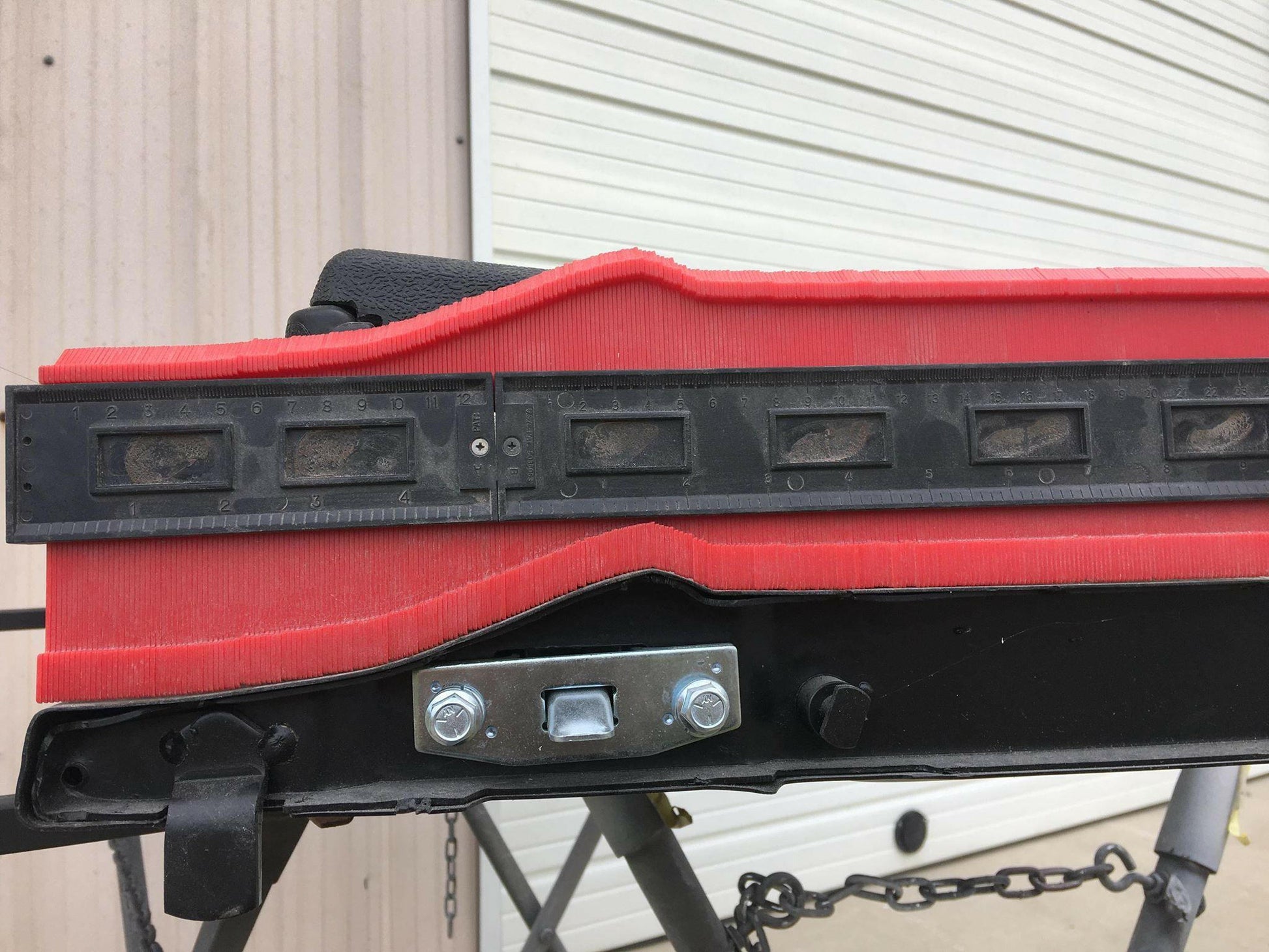 Close-up of the 9livesracing C10 1967-72 Truck Spoiler Kit, featuring a red component with a black adjustable ruler made from durable 6061 aluminum, all placed atop a metal stand. The background shows a corrugated metal wall and a closed garage door. Bolts and chains are attached to the lower section of the kit.
