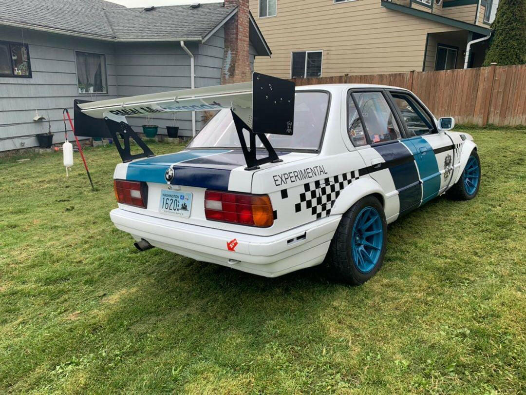 A white BMW E30 with blue accents is parked on grass in a residential area. The car is equipped with a 9livesracing 325 Big Wang kit, providing significant downforce, and features blue alloy wheels along with "EXPERIMENTAL" text on the side. The house in the background has gray siding and brown shingles. A white fire extinguisher is nearby.