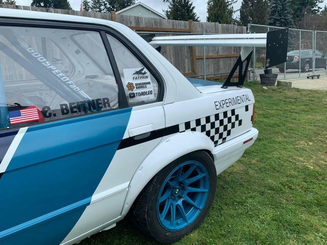 Side view of a modified BMW E30 race car featuring the 9livesracing 325 Big Wang kit ’82-94 E30, with blue accents and "EXPERIMENTAL" emblazoned on the side. The car is equipped with a large rear wing for enhanced downforce, blue wheels, and several stickers—including a small American flag—on the rear window. A wooden fence and grass lawn are visible in the background.