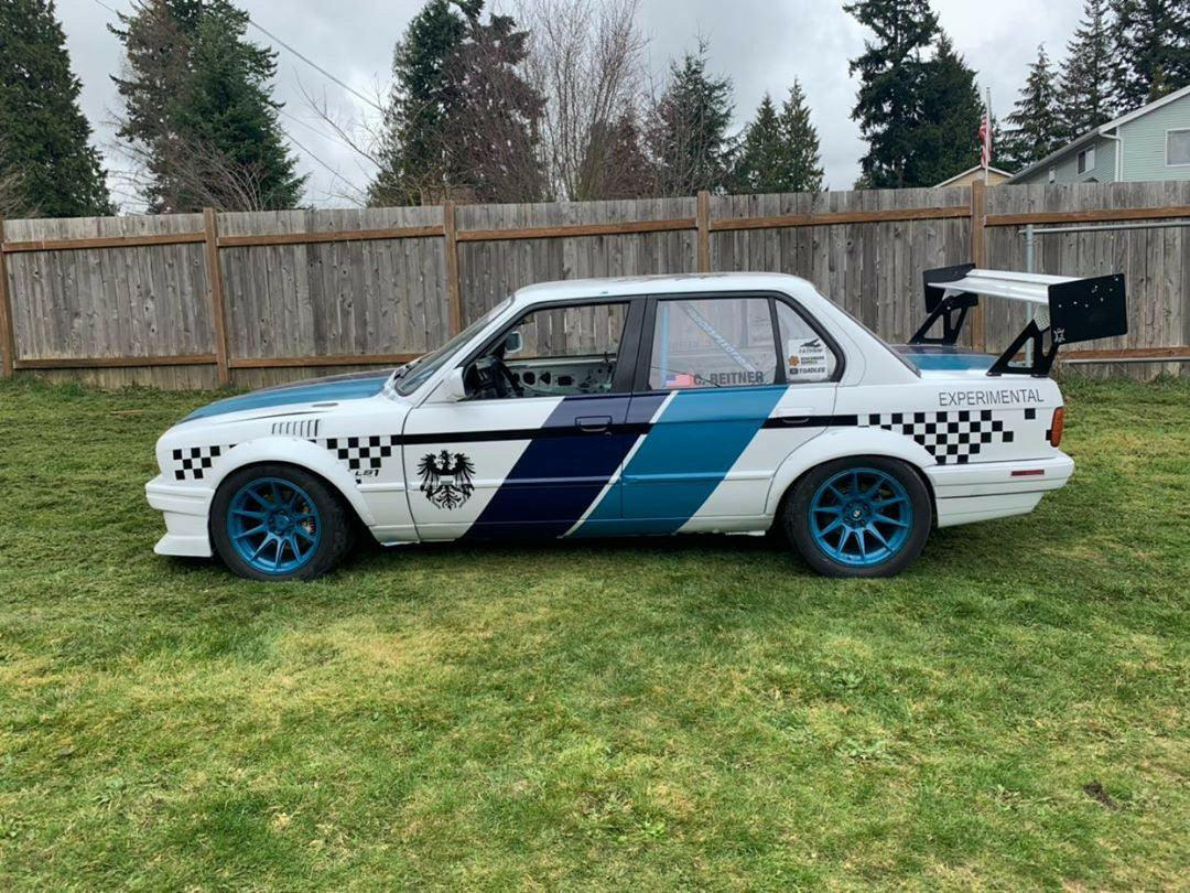 A white sportscar with blue and black checkered patterns, blue wheels, and a large rear wing for added downforce—specifically the 325 Big Wang kit ’82-94 E30 by 9livesracing—is parked on a grassy lawn. The car has "EXPERIMENTAL" written on the side and features a black emblem on the door. A wooden fence and trees are in the background.