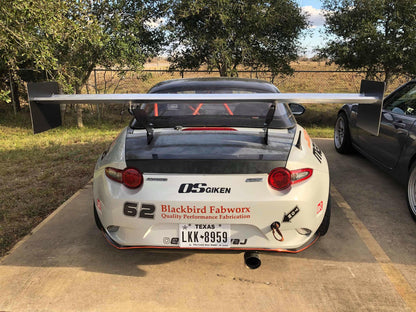 A white sports car with a 9livesracing Miata Big Chassis mount Wang Kit 2016+ ND, adorned with multiple decals including "OS Giken," "Blackbird Fabworx," "62," and a Texas license plate reading "LKN-8959." It is parked on a concrete pad with trees and a grassy field in the background.