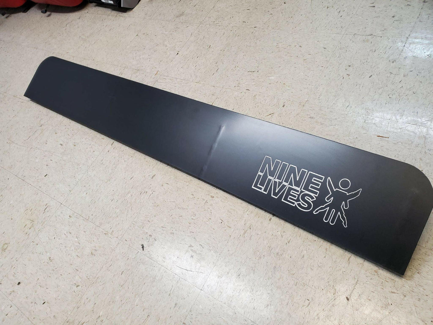 A long, black C10 1967-72 Truck spoiler Kit made from 6061 aluminum with a matte finish is positioned diagonally on a beige tiled floor. The spoiler, designed to enhance downforce, features the text "9livesracing" in white, accompanied by a silhouette of a person with outstretched arms.