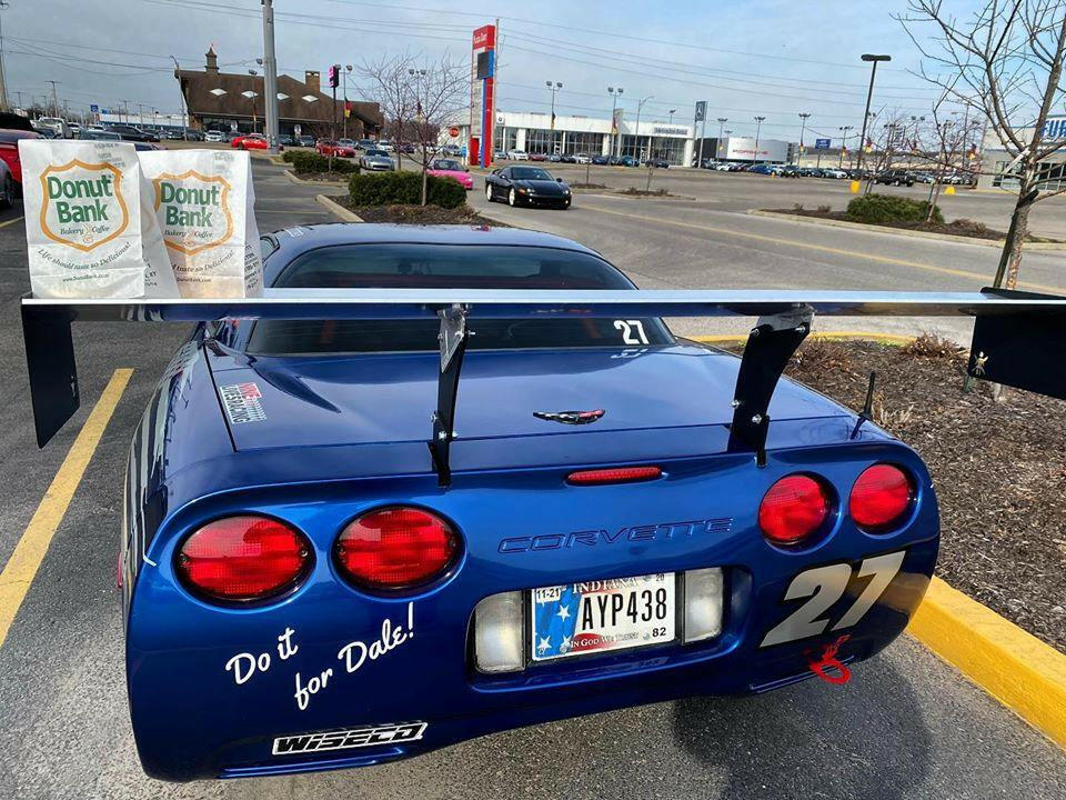 A blue '97-04 C5 Corvette with the number 27 is equipped with a 9livesracing Corvette Medium Downforce Kit and has "Do it for Dale!" written on the bumper. It is parked in a lot with two bags from Donut Bank resting on its rear window. In the background, cars are parked near commercial buildings.