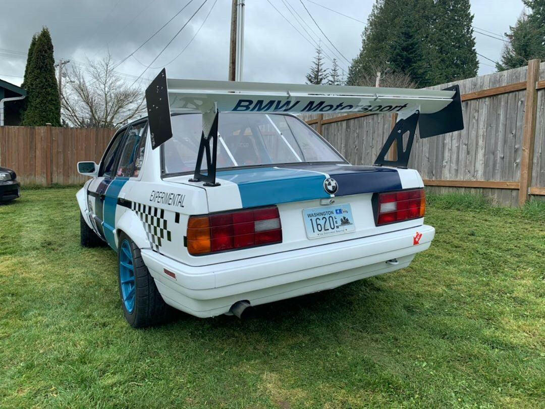 A white custom E30 BMW race car, featuring the 325 Big Wang kit ’82-94 E30 from 9livesracing, is parked on grass. It boasts a substantial rear wing for added downforce, blue wheels, and black-and-white checkered decals on its sides. The car's license plate reads "1620X," and it is adorned with the "BMW Motorsport" text on the Big Wang. In the background, there are a wooden fence and trees.