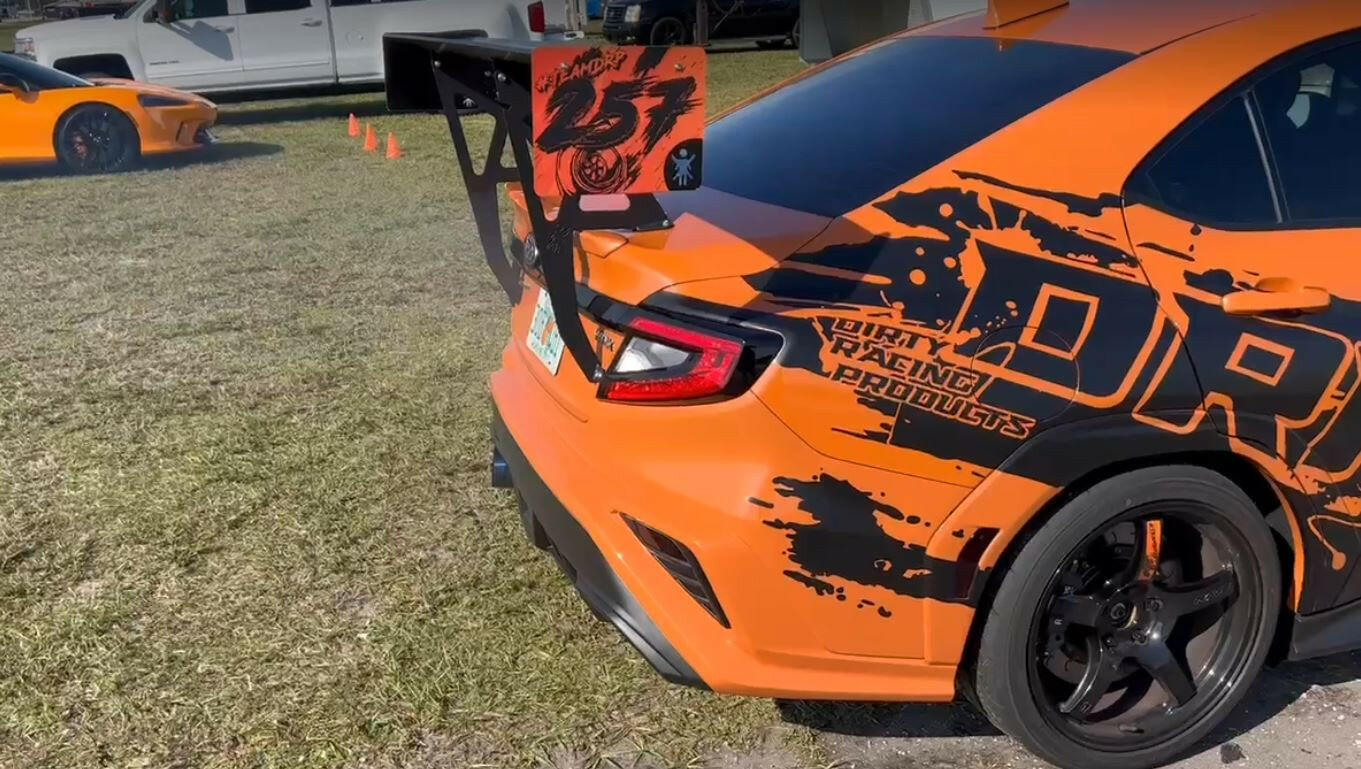 A Subaru Impreza STI with the 9livesracing Big Wang kit '2022 - current edition is on display in a grassy area. The orange sports car boasts a large black rear spoiler and features an eye-catching black and orange graphic design with the text "Drift Racing Products." Downforce adjustments ensure optimal performance. In the background, another orange sports car can be seen along with a white truck.