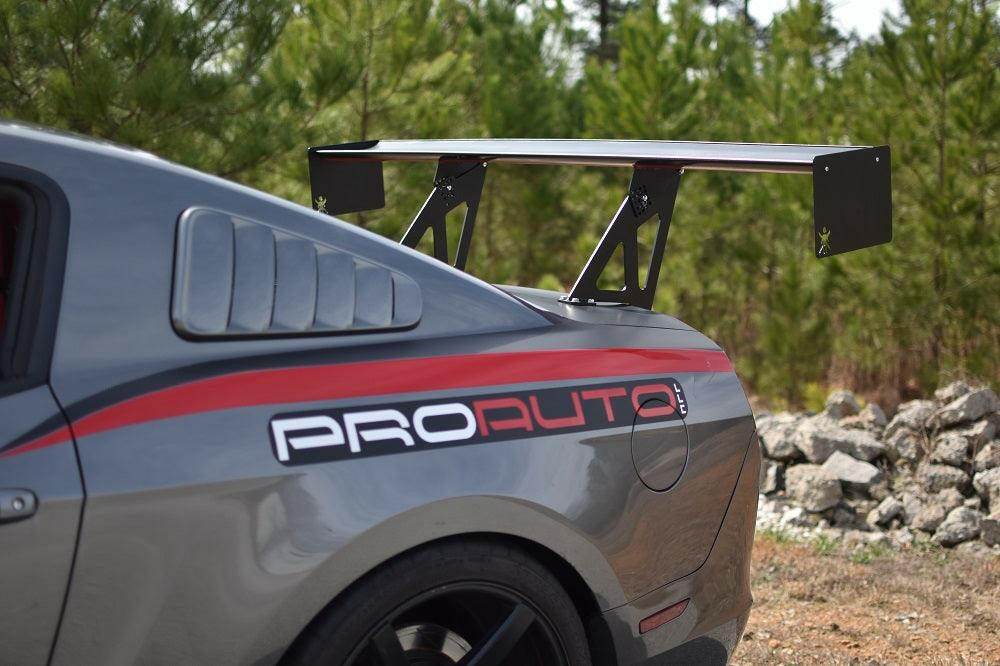 Close-up of a gray sports car's rear, featuring the 9livesracing Mustang Big Wang Kit '10-14 S197 II with adjustable downforce, black alloy wheels, and a "PROAUTO" decal on the side. The background shows green trees and a rocky area.