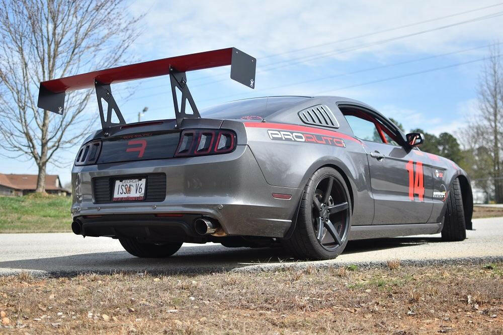 A sleek gray sports car with a large rear wing and black alloy wheels is parked on a road. Equipped with the Mustang Big Wang Kit '10-14 S197 II by 9livesracing, which includes extruded aluminum wings for downforce adjustment, it prominently features a racing number "14" on the side and the text "PROAUTO" on the rear window. Trees and a house can be seen in the background under a partly cloudy sky.