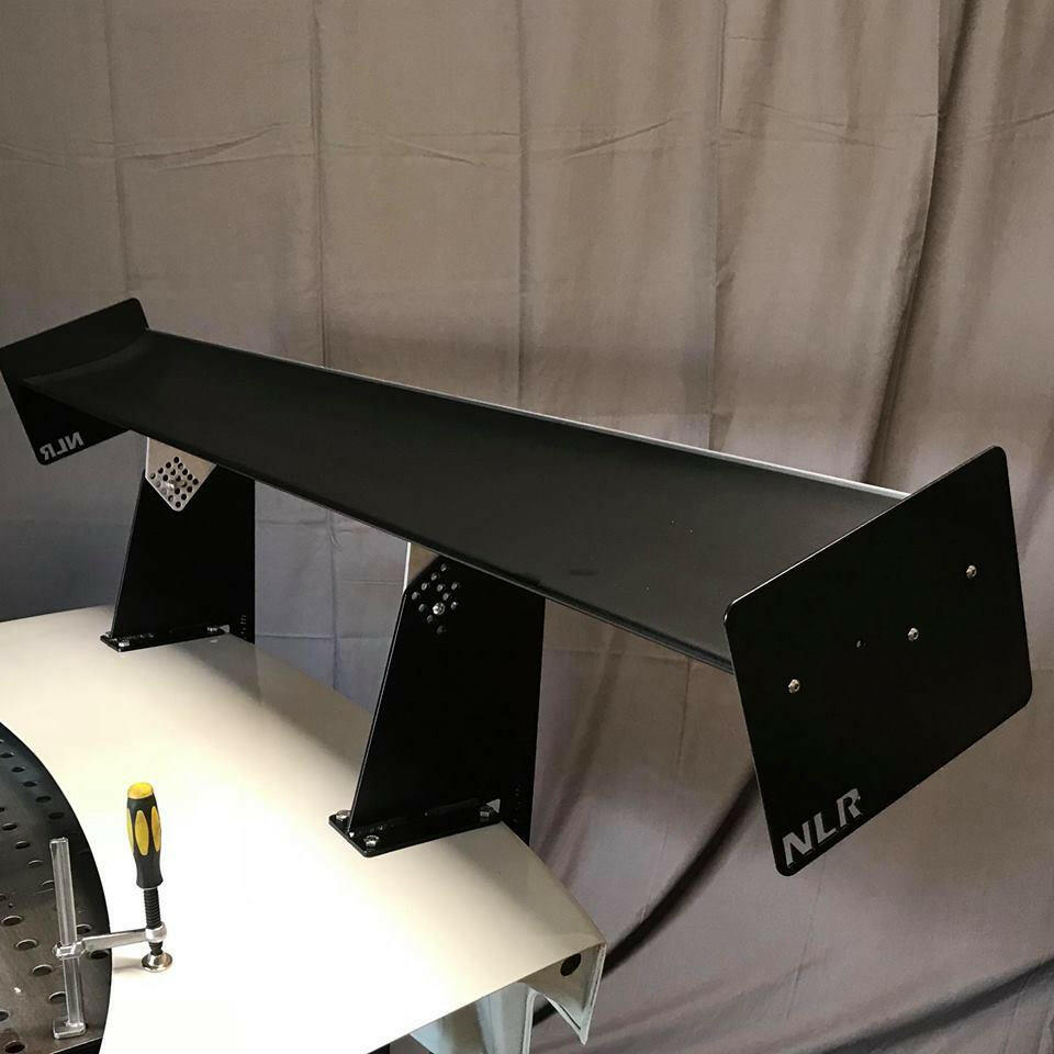 A black metal wing spoiler with "NLR" branding is mounted on a white desk with tool holes, using the BMW E36 Mounts/Pylons Only kit and aluminum hardware from 9livesracing. A yellow and black-handled screwdriver rests near one of the mounting brackets. Gray curtains hang in the background.