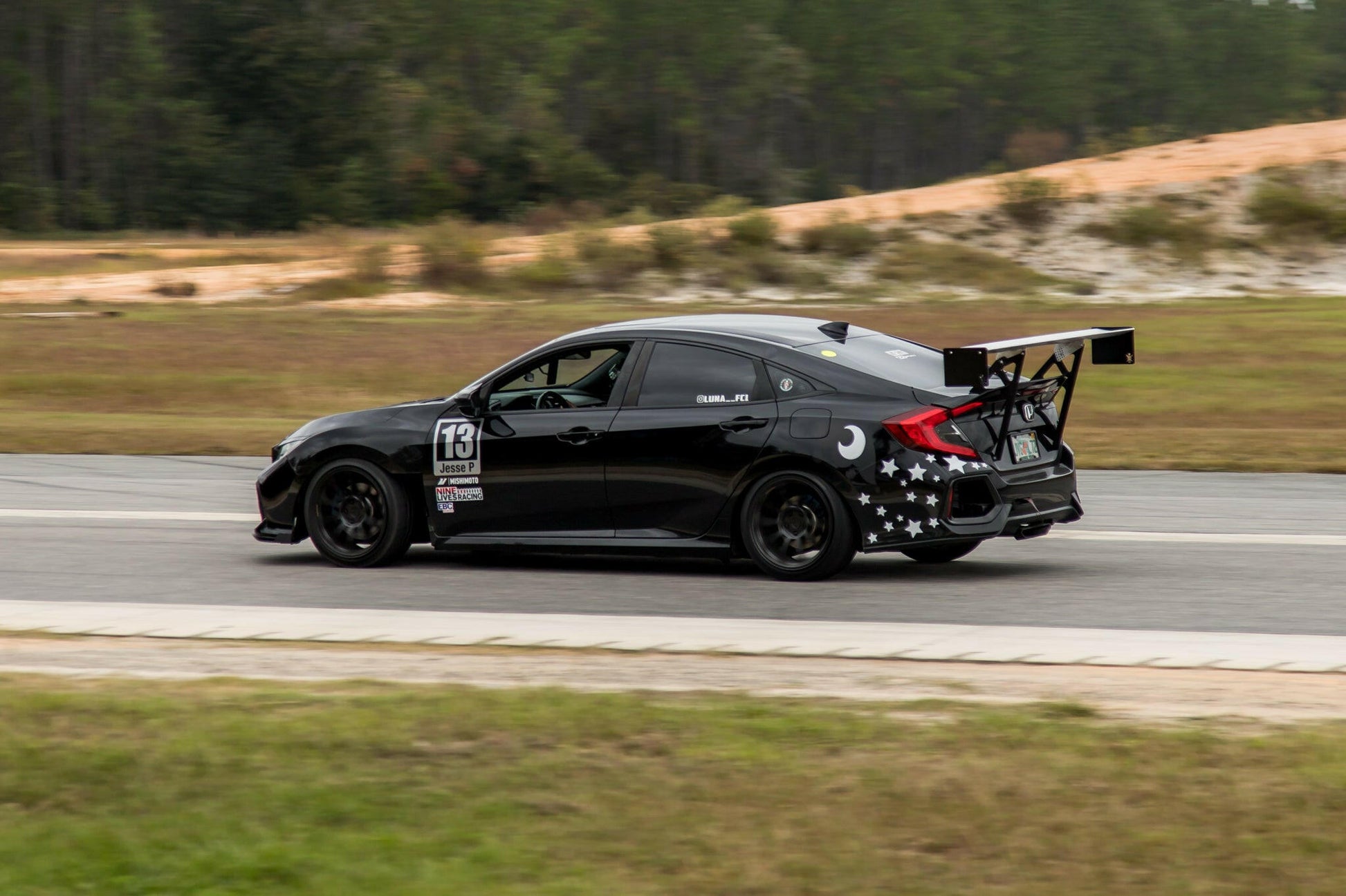 A black sports car with number 13 on its door speeds on a racetrack. The car, equipped with a Civic Wang kit 2017+ FC1 from 9livesracing for optimum downforce, features star and moon decals on the side. The background shows a grassy area and some trees.