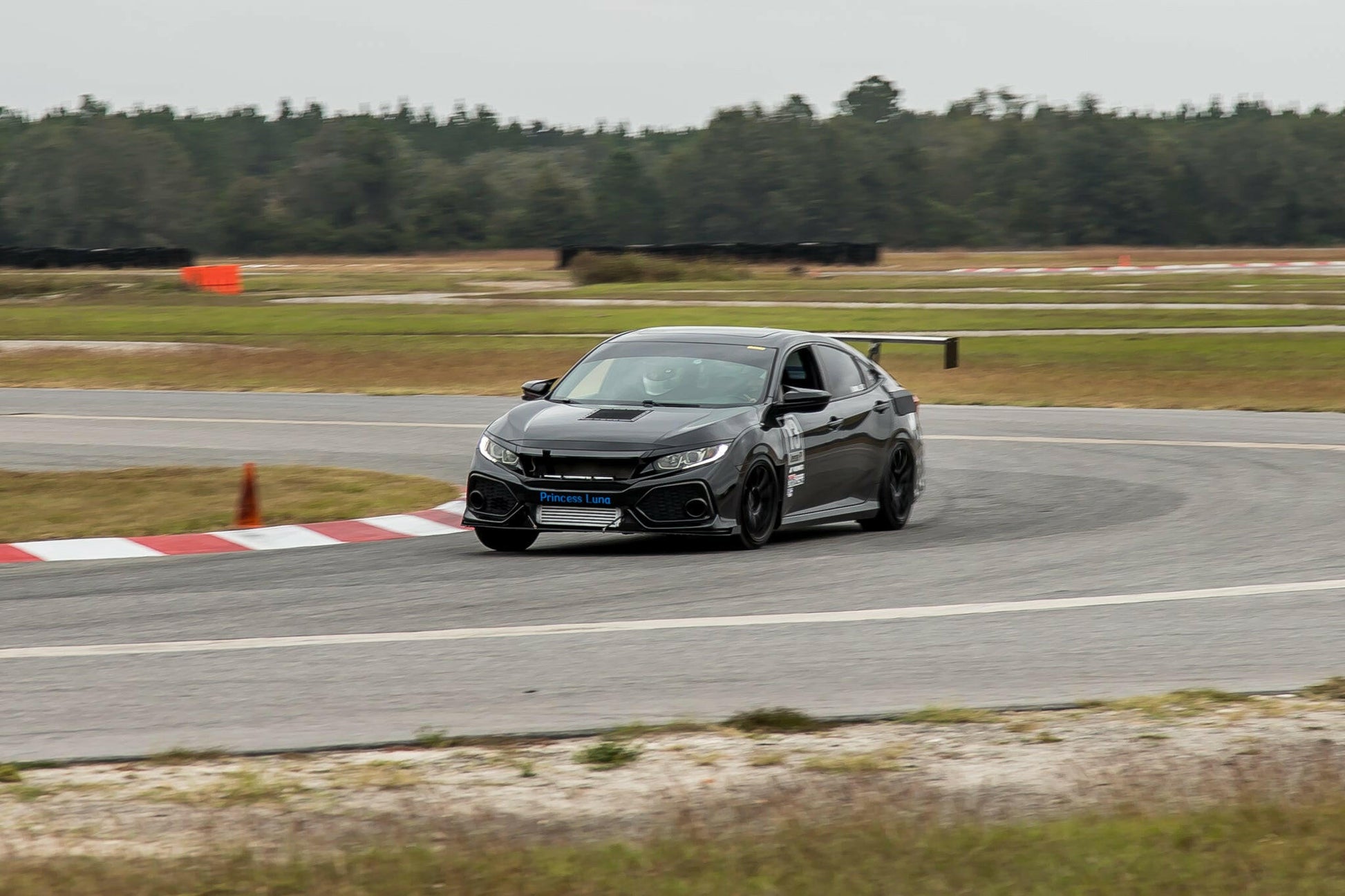 A sleek, black sports car equipped with an impressive Civic Wang kit 2017+ FC1 from 9livesracing speeds around a curve on a racetrack, against a background of greenery and a cloudy sky. The car is positioned close to the red and white striped corner marker.