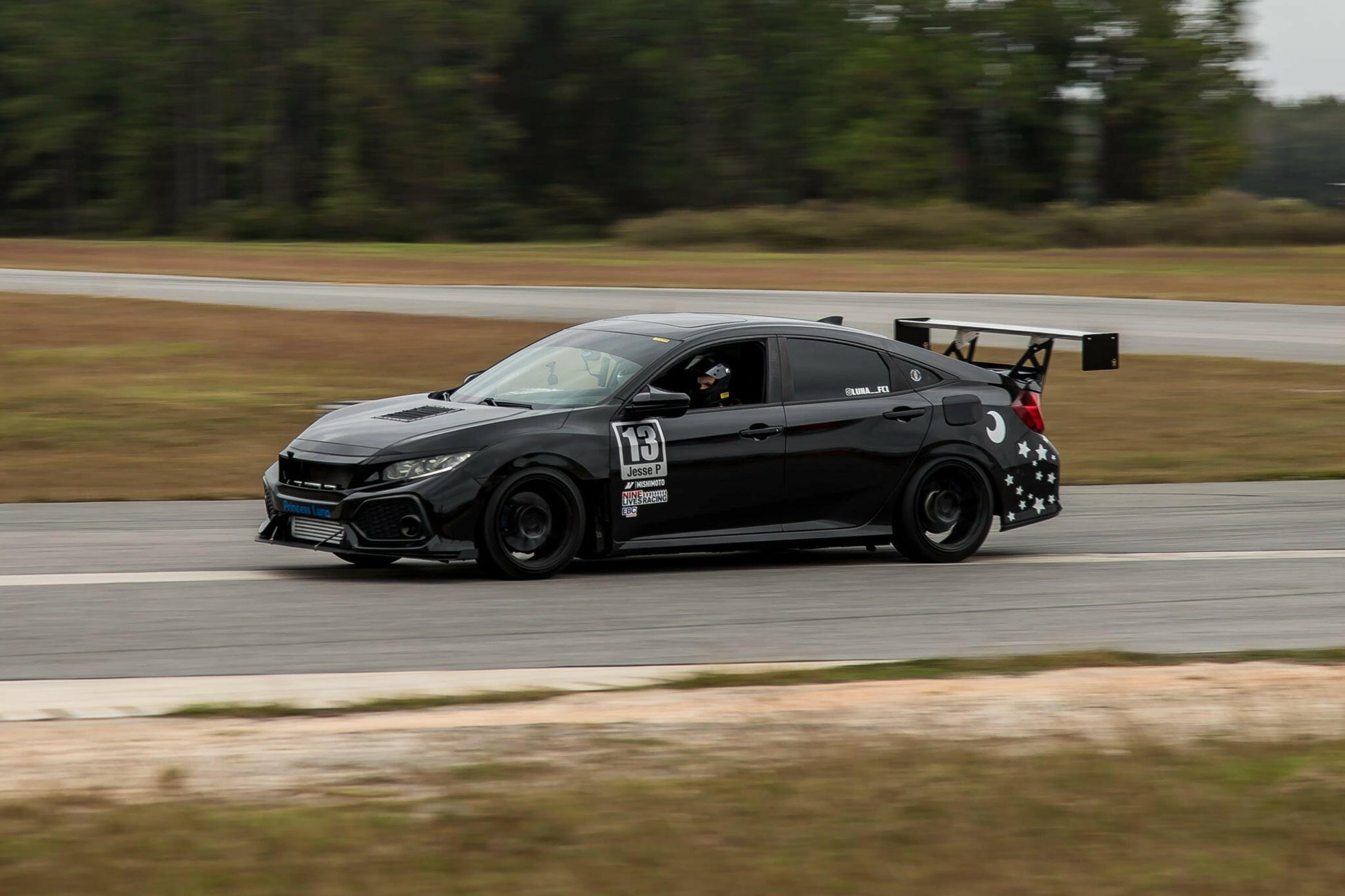 A black, modified race car with the number 19 on the door, equipped with a Big Wang Civic kit 2017+ FC1 rear wing from 9livesracing and adorned with various stickers, speeds down a track against a backdrop of trees and grass. The car's driver, wearing a helmet, is visible through the window as aluminum wings generate exceptional downforce.