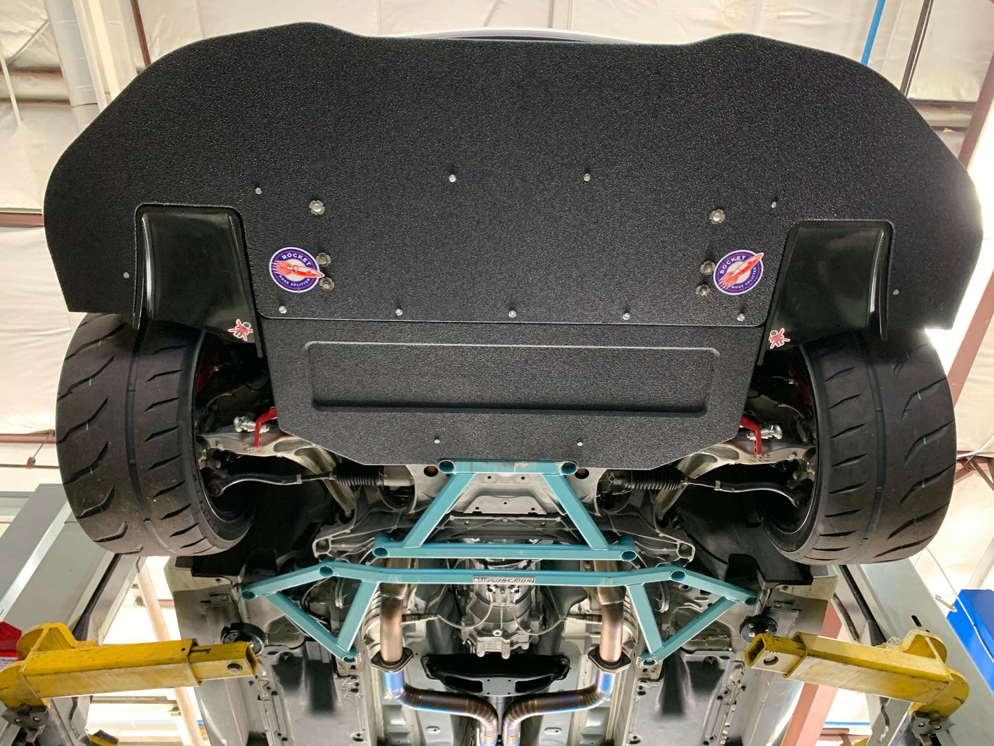 View from underneath a 370z Rocket Nose v2 Splitter ’08-20 Z34, by 9livesracing, showing the rear undercarriage. Visible are large tires, suspension components, exhaust system, and a diffuser with performance stickers enhancing downforce. The garage ceiling and hydraulic lift arms can also be seen.