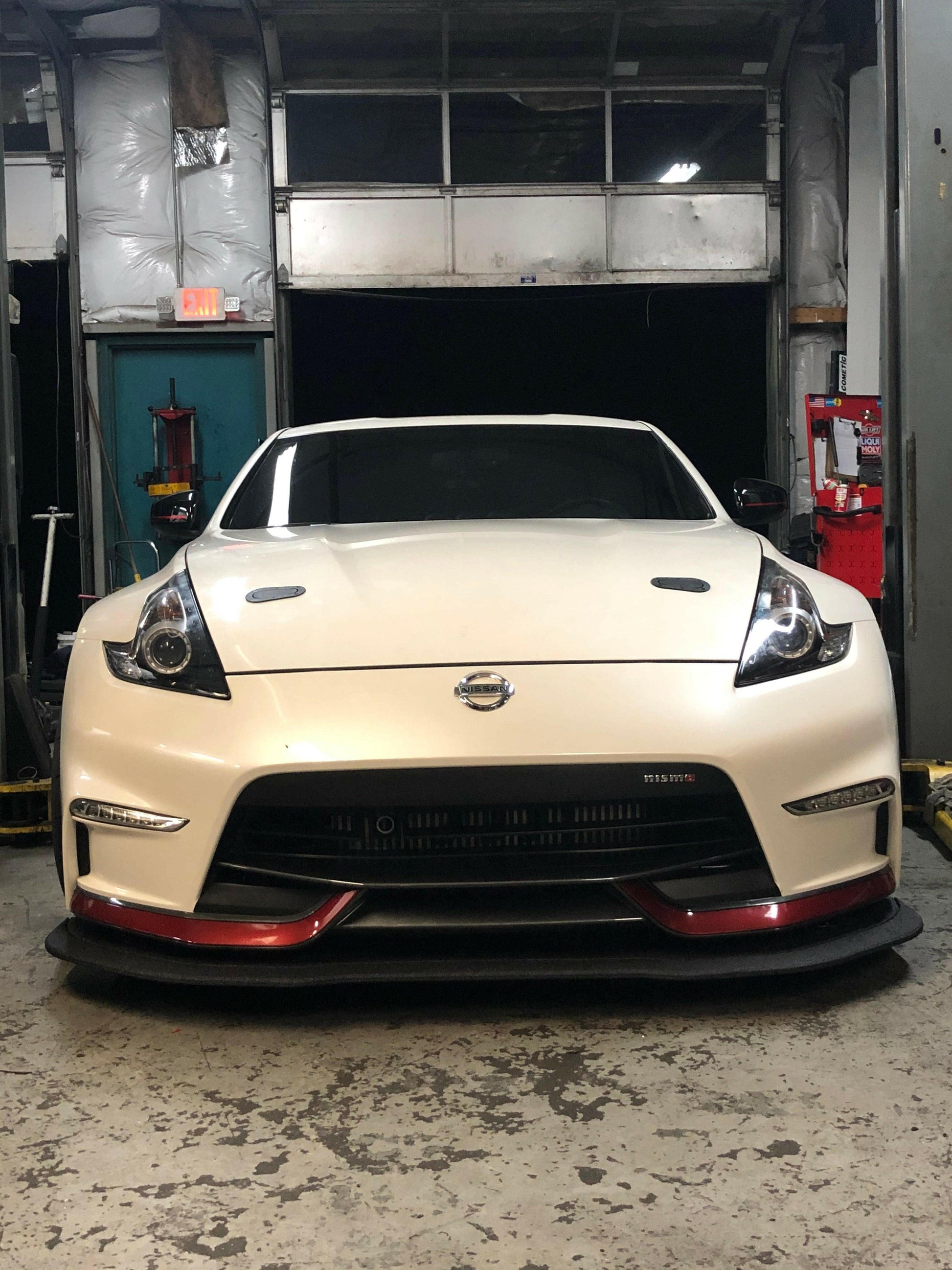 A white sports car, probably a Nissan 370Z, is parked inside a garage with its front facing the camera. The car showcases sleek headlights, a prominent front grille, and red accents on the lower bumper. A 9livesracing 370z Rocket Nose v2 Splitter ’08-20 Z34 enhances its aerodynamic downforce while various tools and equipment are visible in the background.