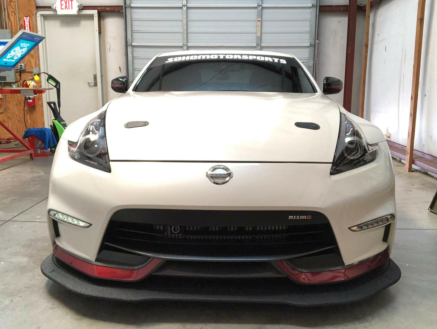 A white sports car branded with Nismo is parked in a garage, proudly showcasing its Rocket Nose Coating and the prominent 370z Rocket Nose v2 Splitter ’08-20 Z34 from 9livesracing. The car features hood vents and a "SONOMOTORSPORTS" banner on the windshield, with a partially visible blue screen-printing press and other workshop equipment in the background.